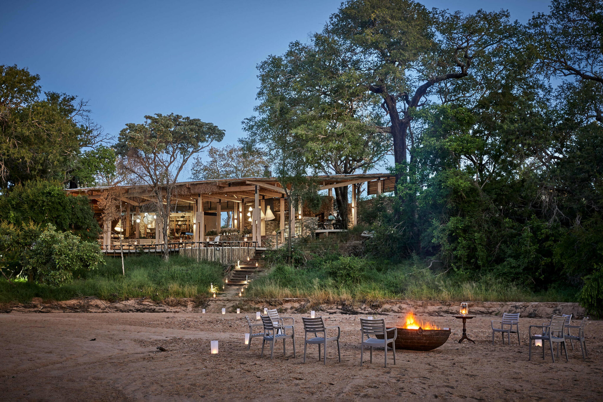 A safari camp lounge and firepit sit by a sandy riverbed at dusk, with chairs arranged under tall trees nearby.