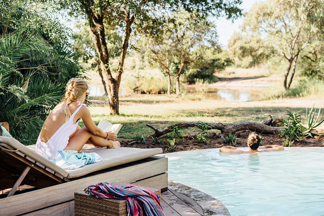 One guest lounges on a chair beside the pool while another floats in the water, with trees and sand beyond.