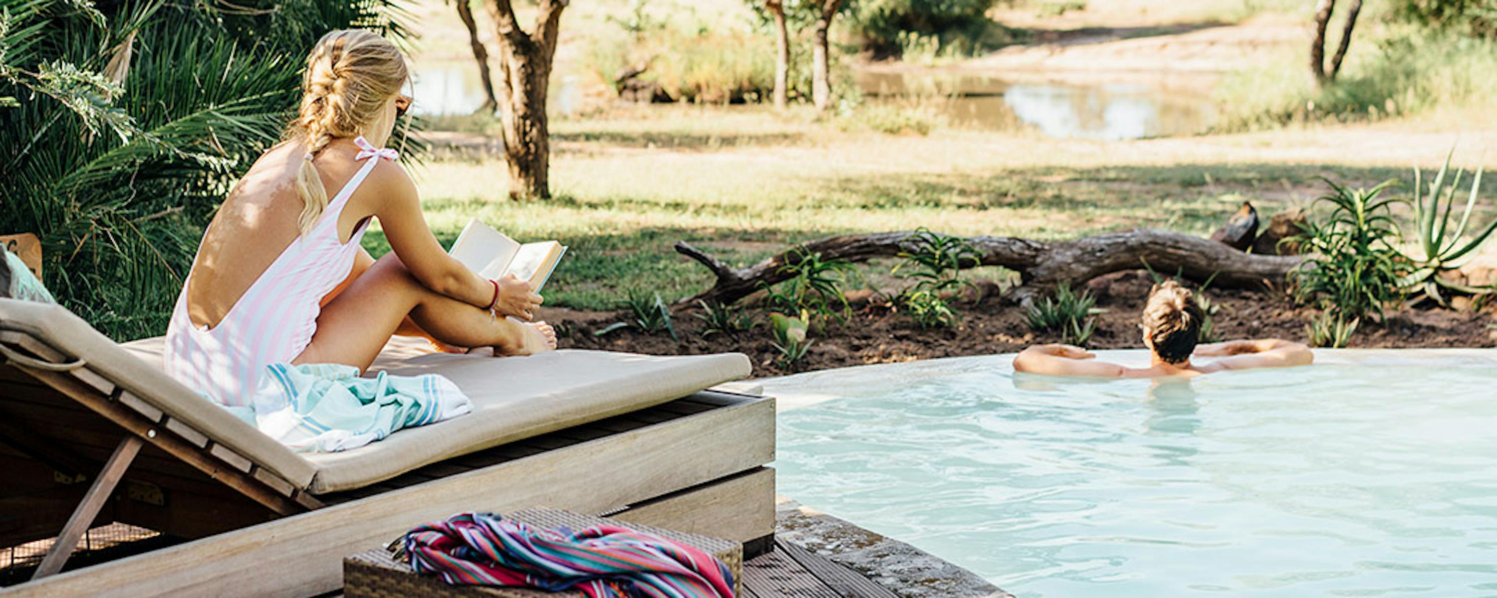 One guest lounges on a chair beside the pool while another floats in the water, with trees and sand beyond.