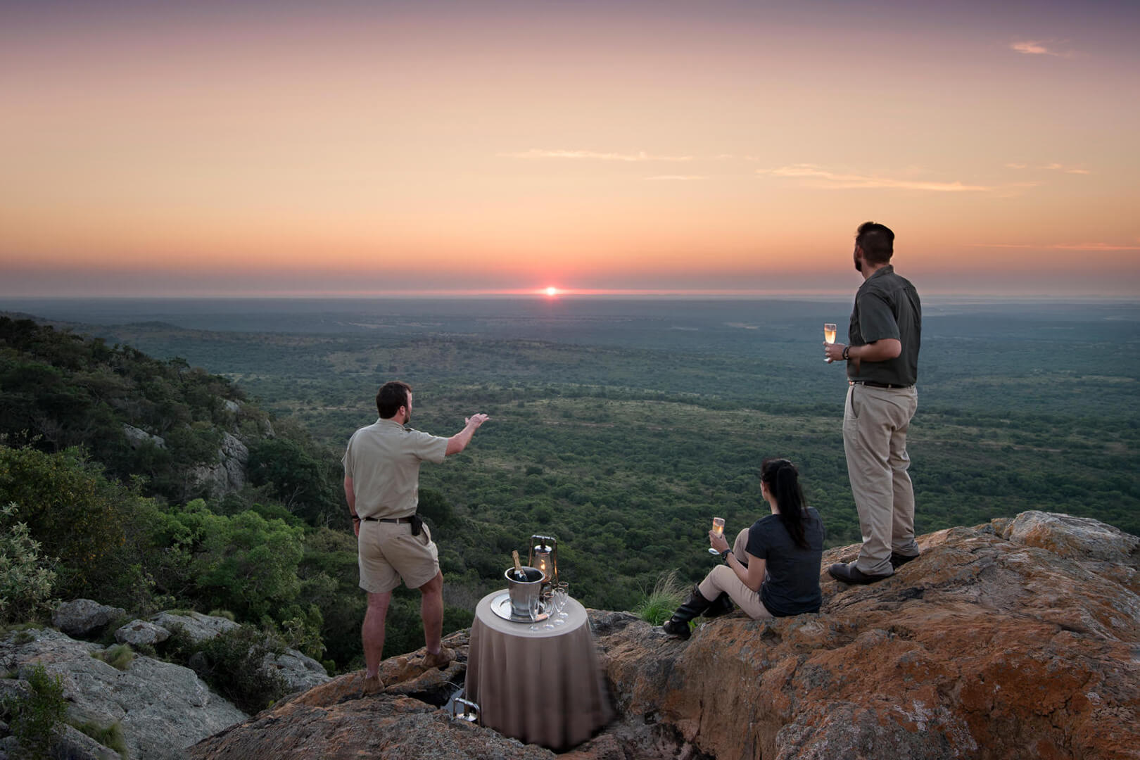Three guests share drinks on a rocky ledge at sunset, looking out over treetops and distant hills far below.