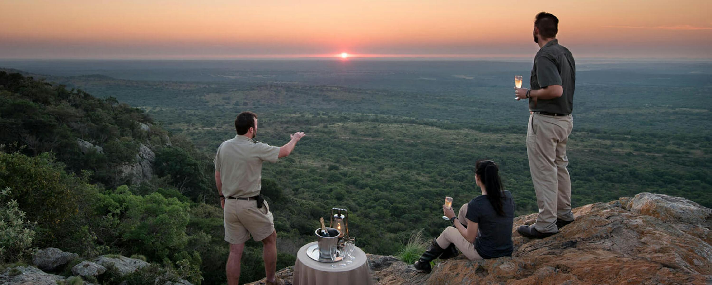 Three guests share drinks on a rocky ledge at sunset, looking out over treetops and distant hills far below.