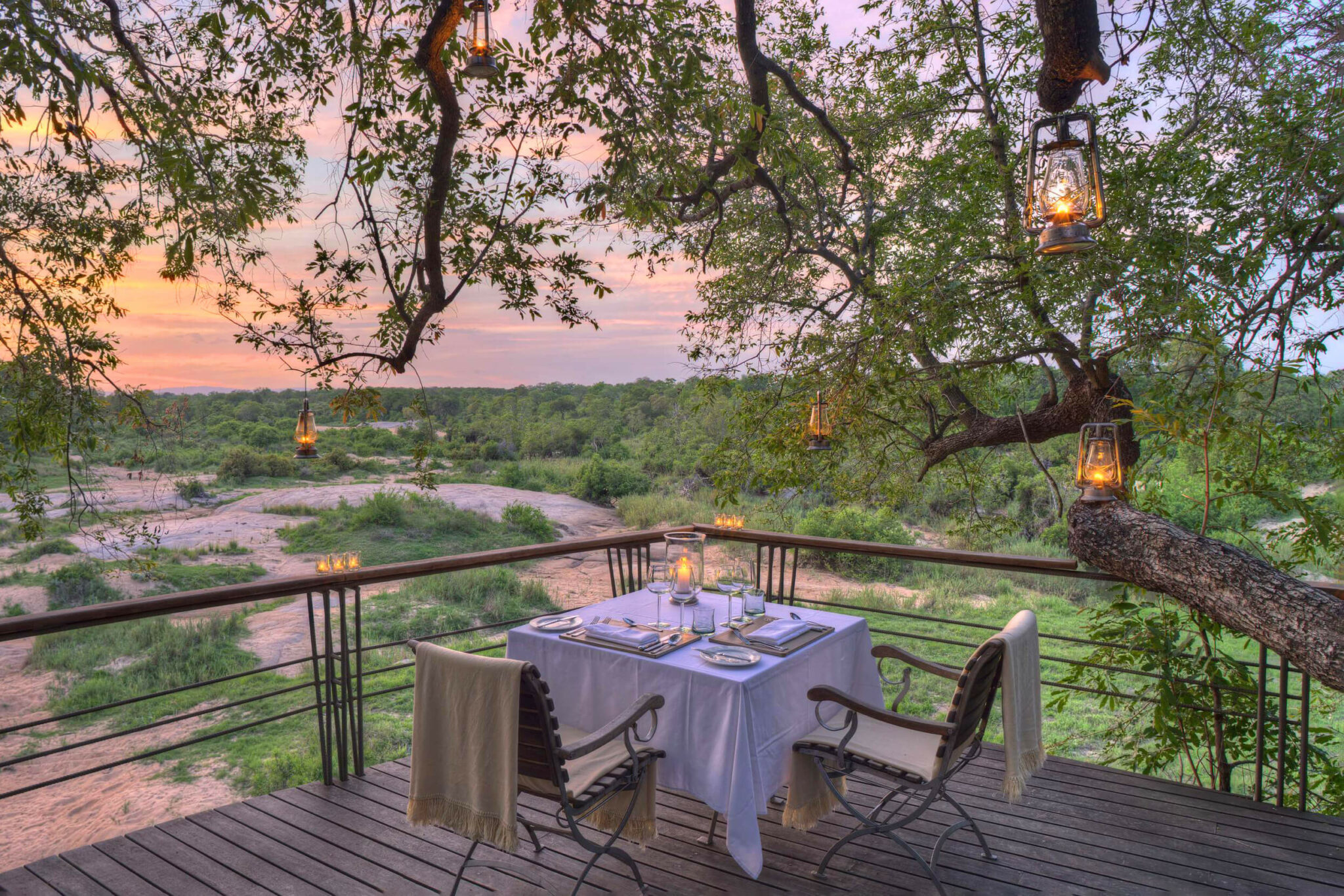 A set dinner table sits on a wooden deck at sunset, with lanterns in branches and the bush beyond the railing.