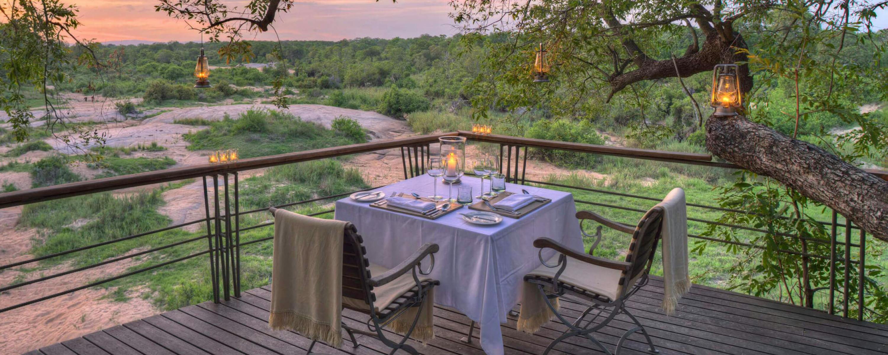A set dinner table sits on a wooden deck at sunset, with lanterns in branches and the bush beyond the railing.