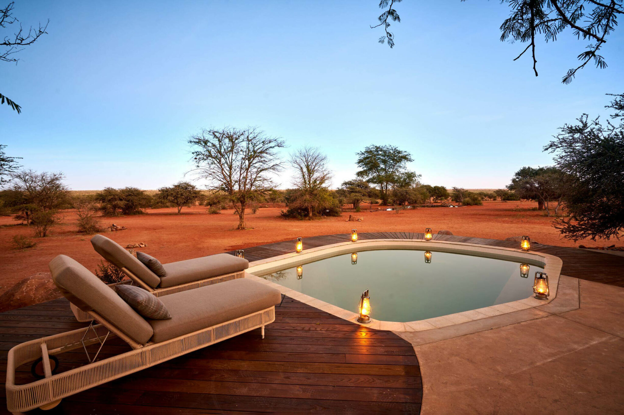 A small plunge pool and cushioned loungers sit on a deck in red desert scrub, softly lit by lanterns at twilight.