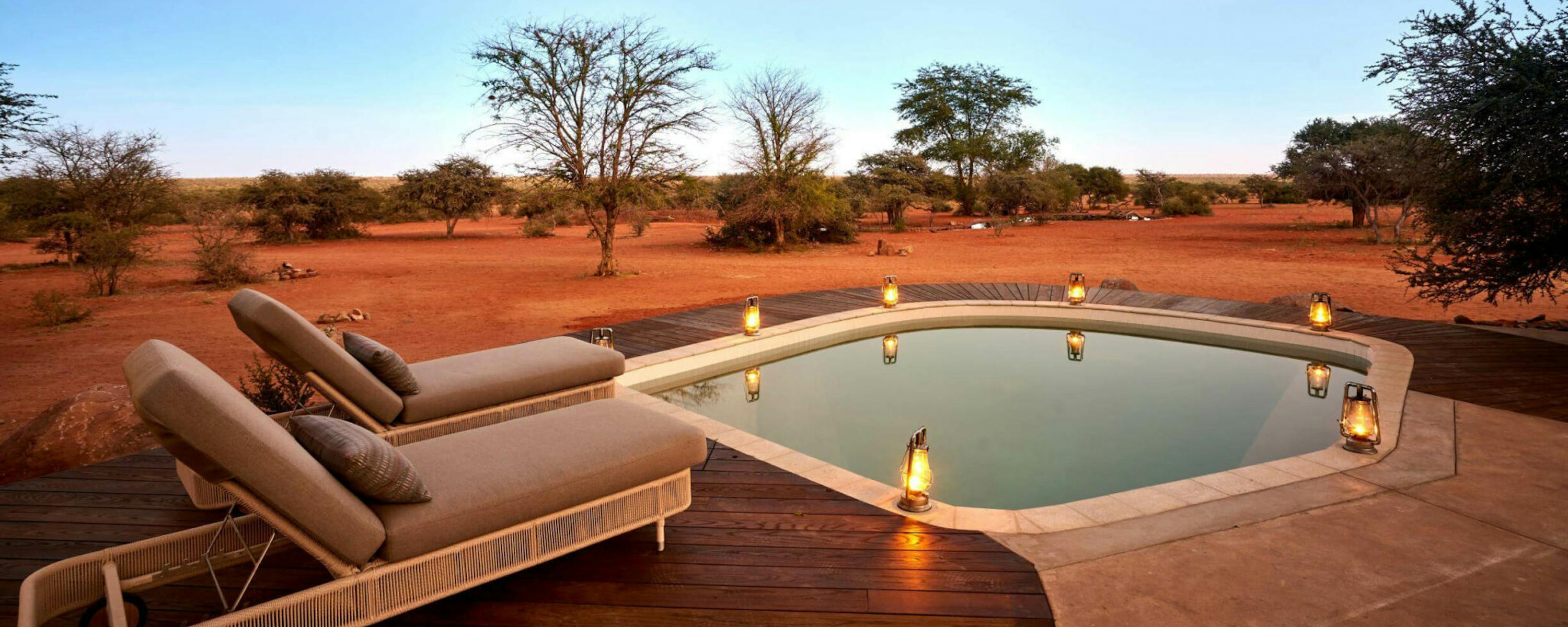 A small plunge pool and cushioned loungers sit on a deck in red desert scrub, softly lit by lanterns at twilight.