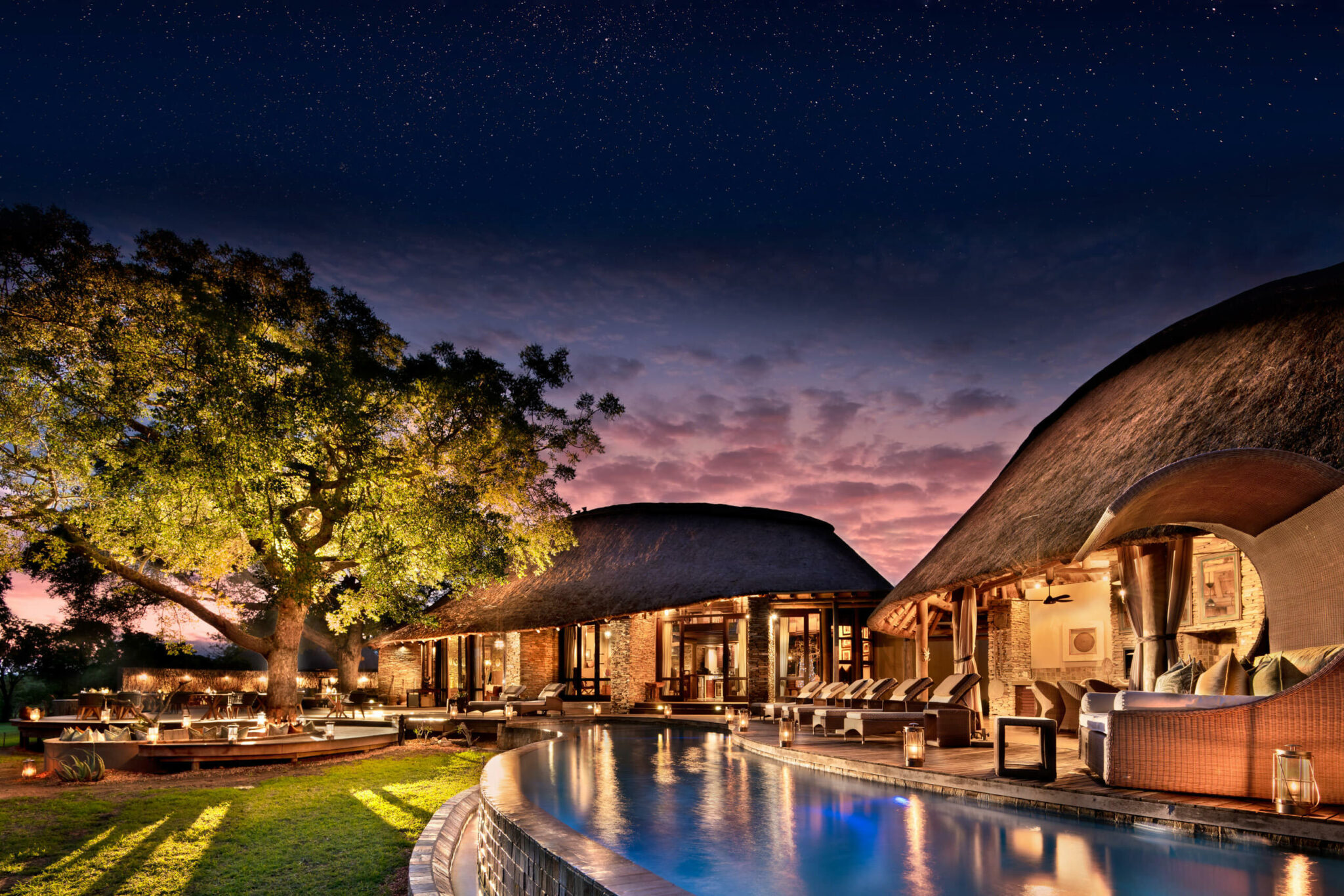 A thatched lodge and curving pool glow at dusk, with lantern-lit seating and trees silhouetted in blue light.