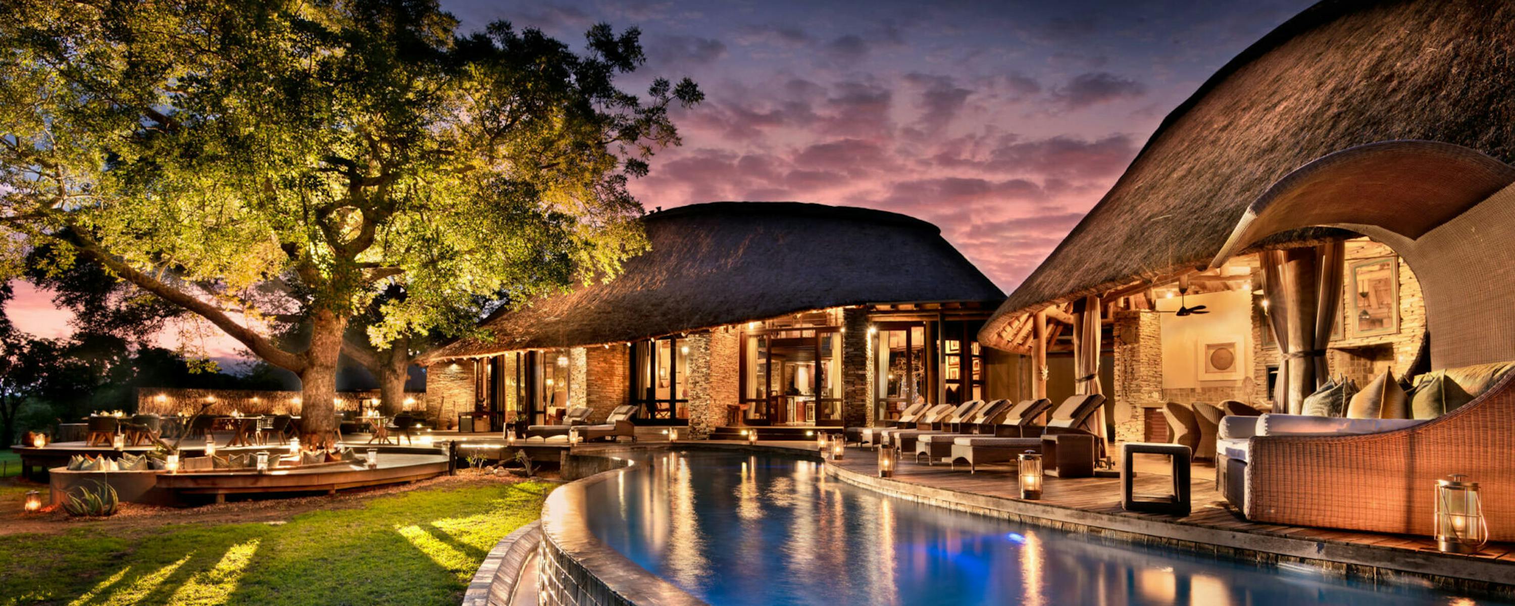 A thatched lodge and curving pool glow at dusk, with lantern-lit seating and trees silhouetted in blue light.