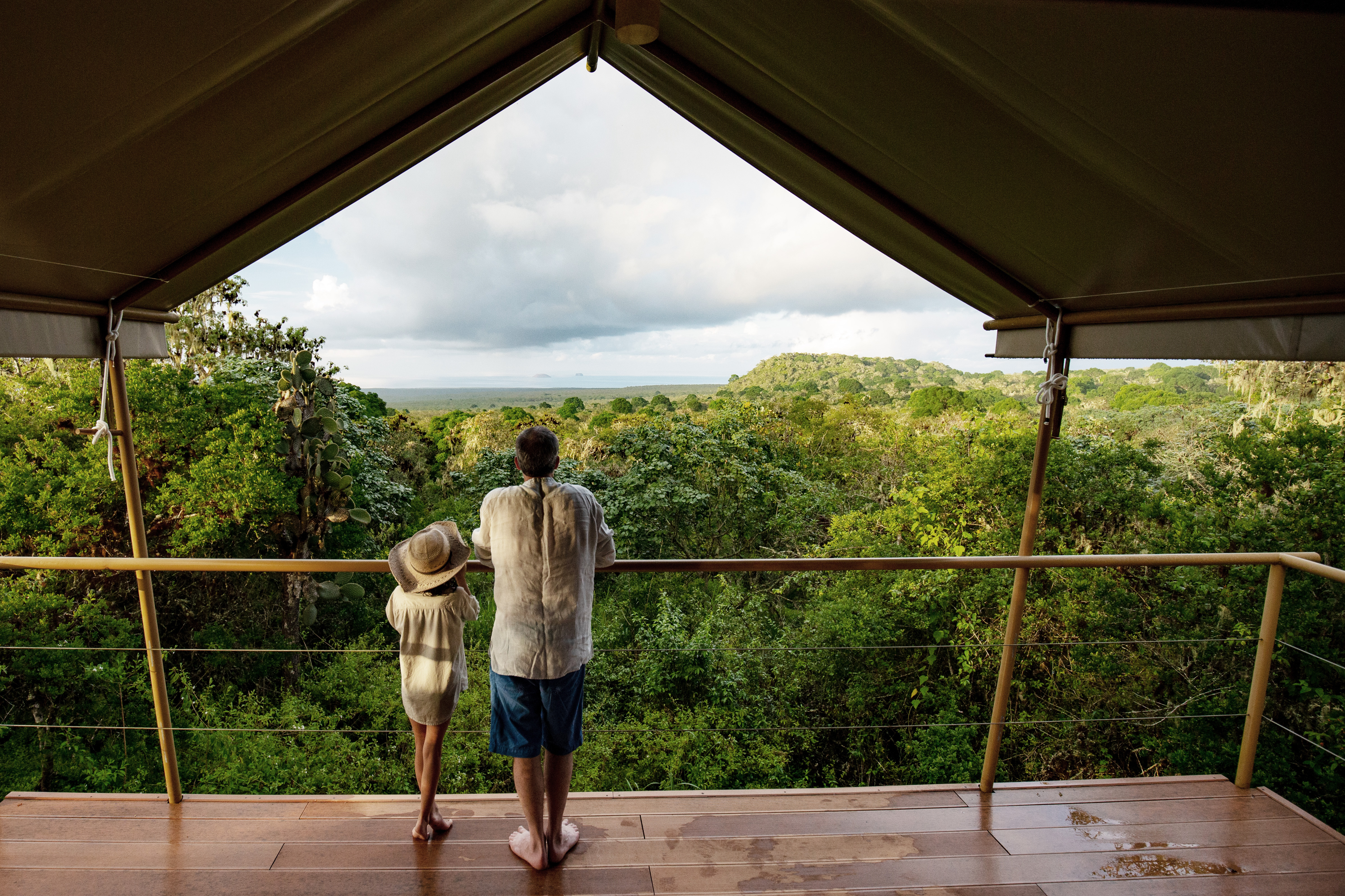 Two guests stand on a covered deck at a safari camp, looking over forested hills beneath cool, cloudy skies.