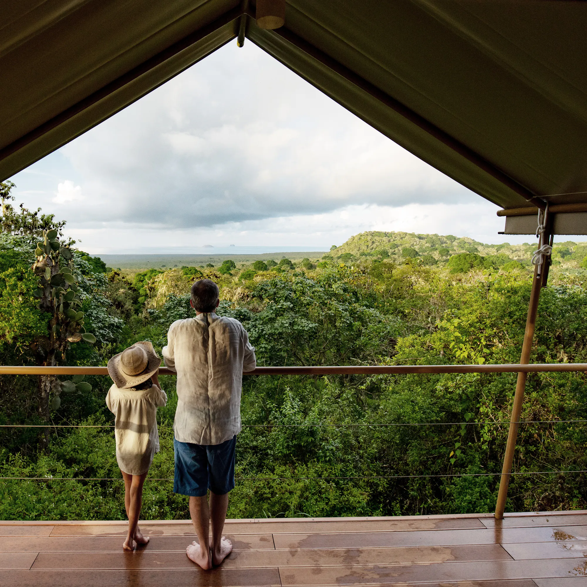 Two guests stand on a covered deck at a safari camp, looking over forested hills beneath cool, cloudy skies.