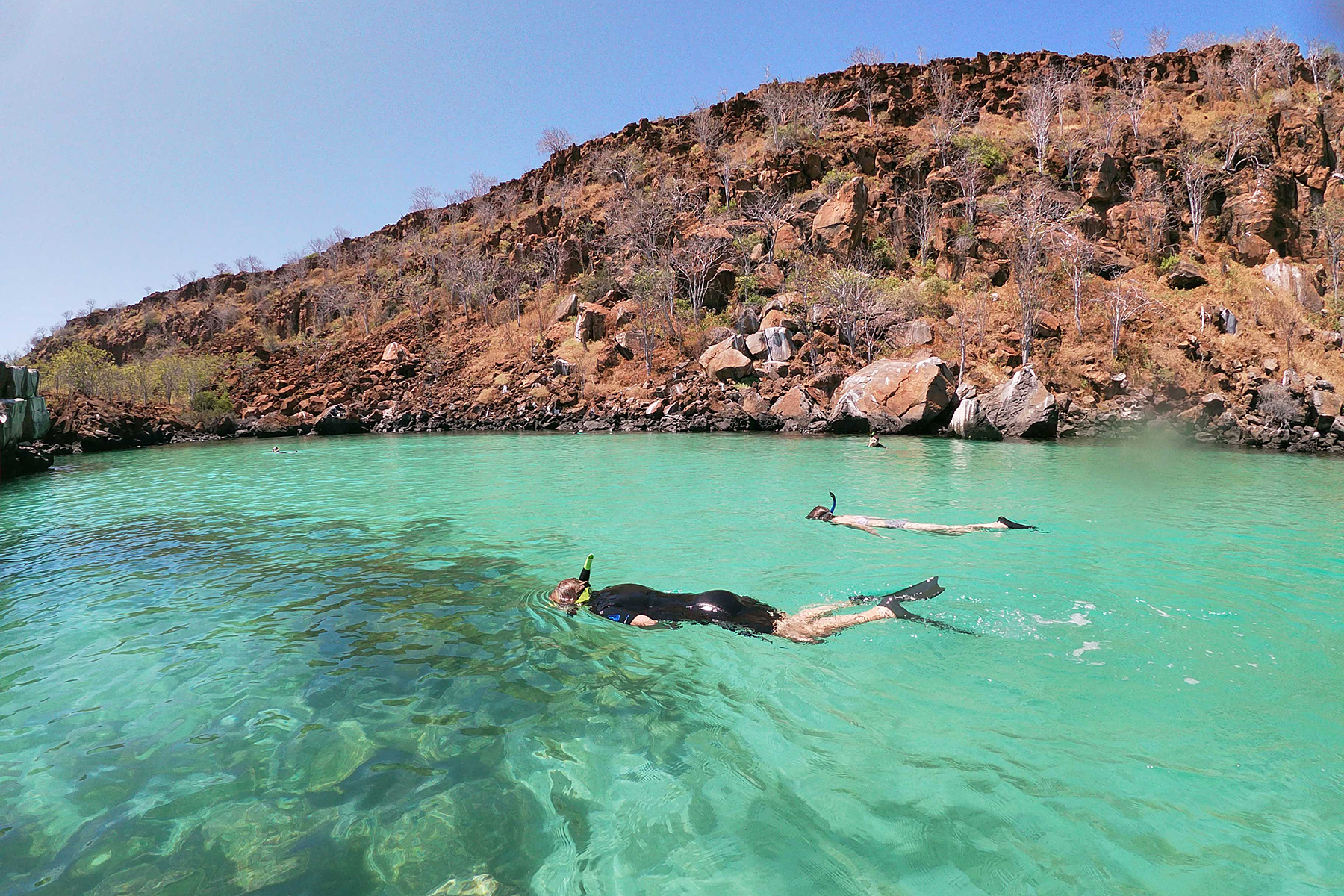 Two swimmers snorkel in clear turquoise water near a rocky shoreline, sunlit shallows revealing the seabed.