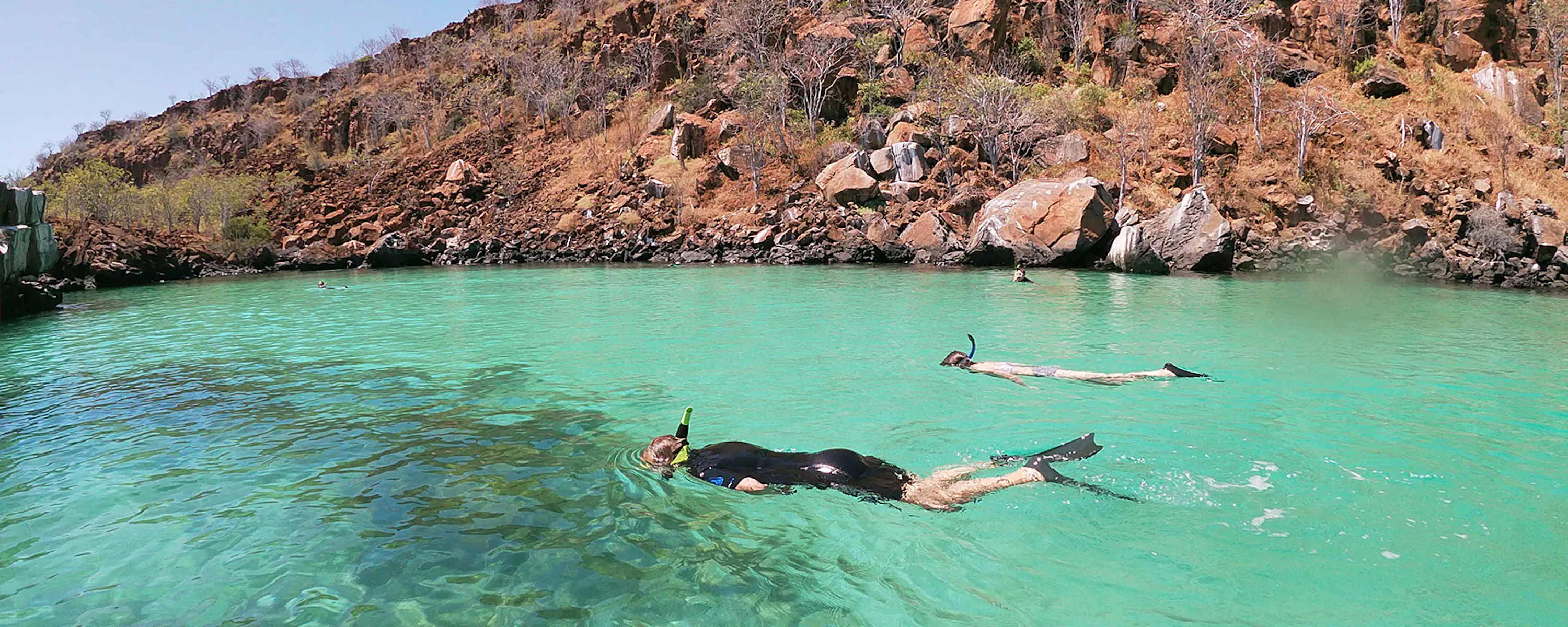 Two swimmers snorkel in clear turquoise water near a rocky shoreline, sunlit shallows revealing the seabed.
