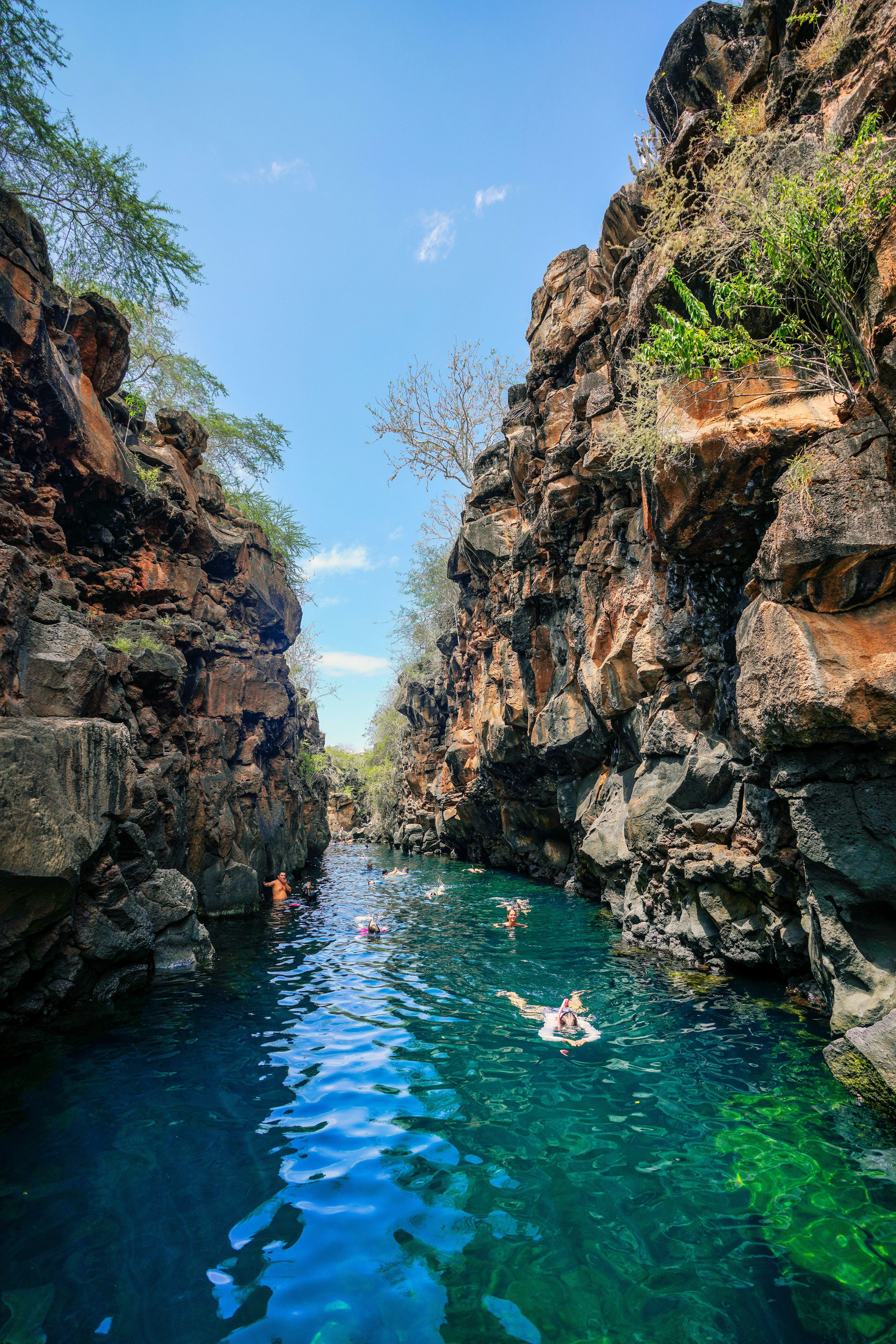 Swimmers float in a narrow rock canyon, sunlight reflecting on bright blue water between steep cliff walls.