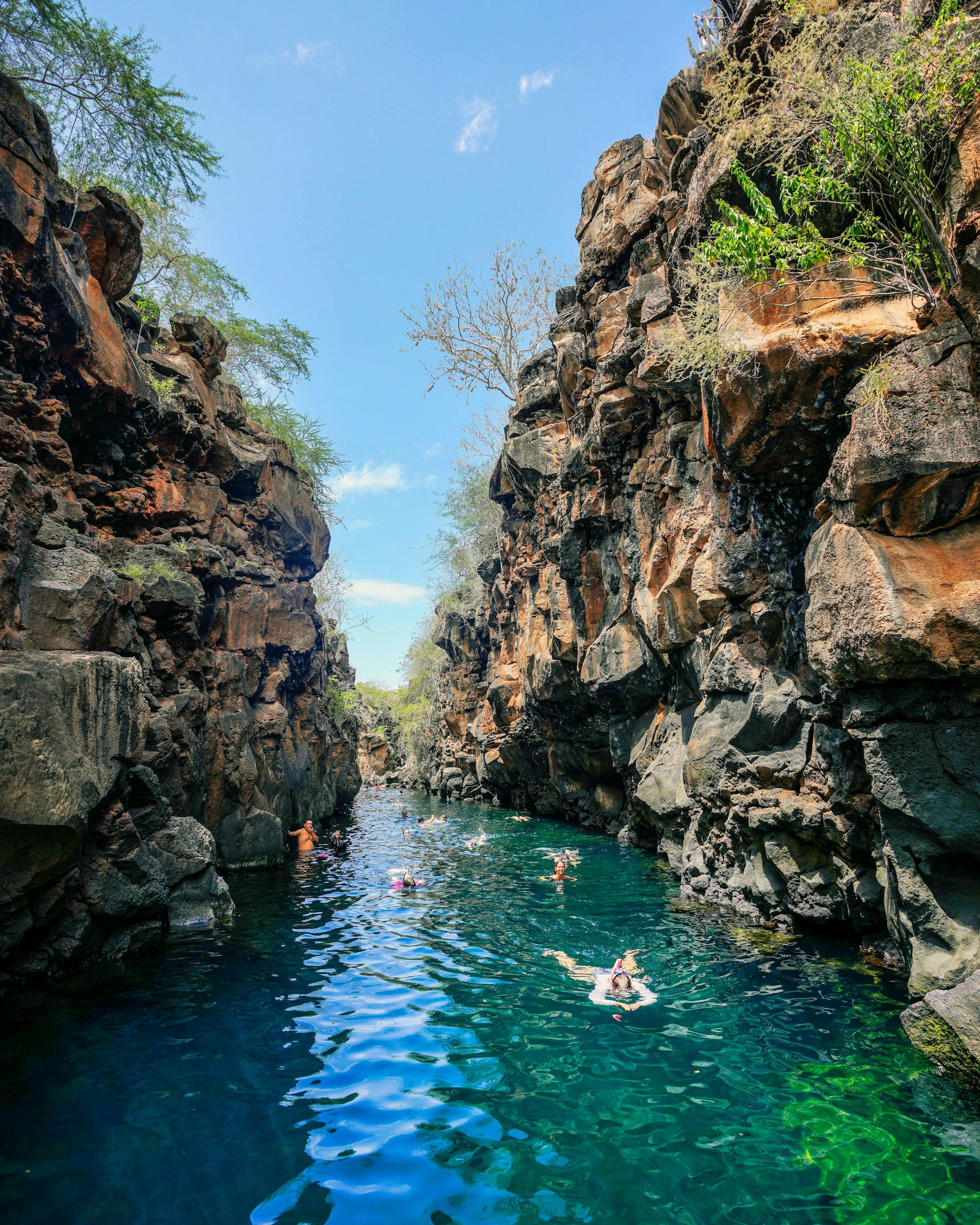 Swimmers float in a narrow rock canyon, sunlight reflecting on bright blue water between steep cliff walls.