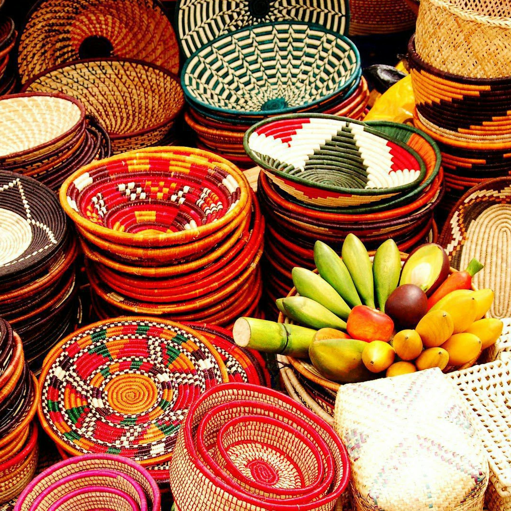 Brightly patterned woven baskets are stacked at a market stall, with bananas and citrus fruit piled nearby.