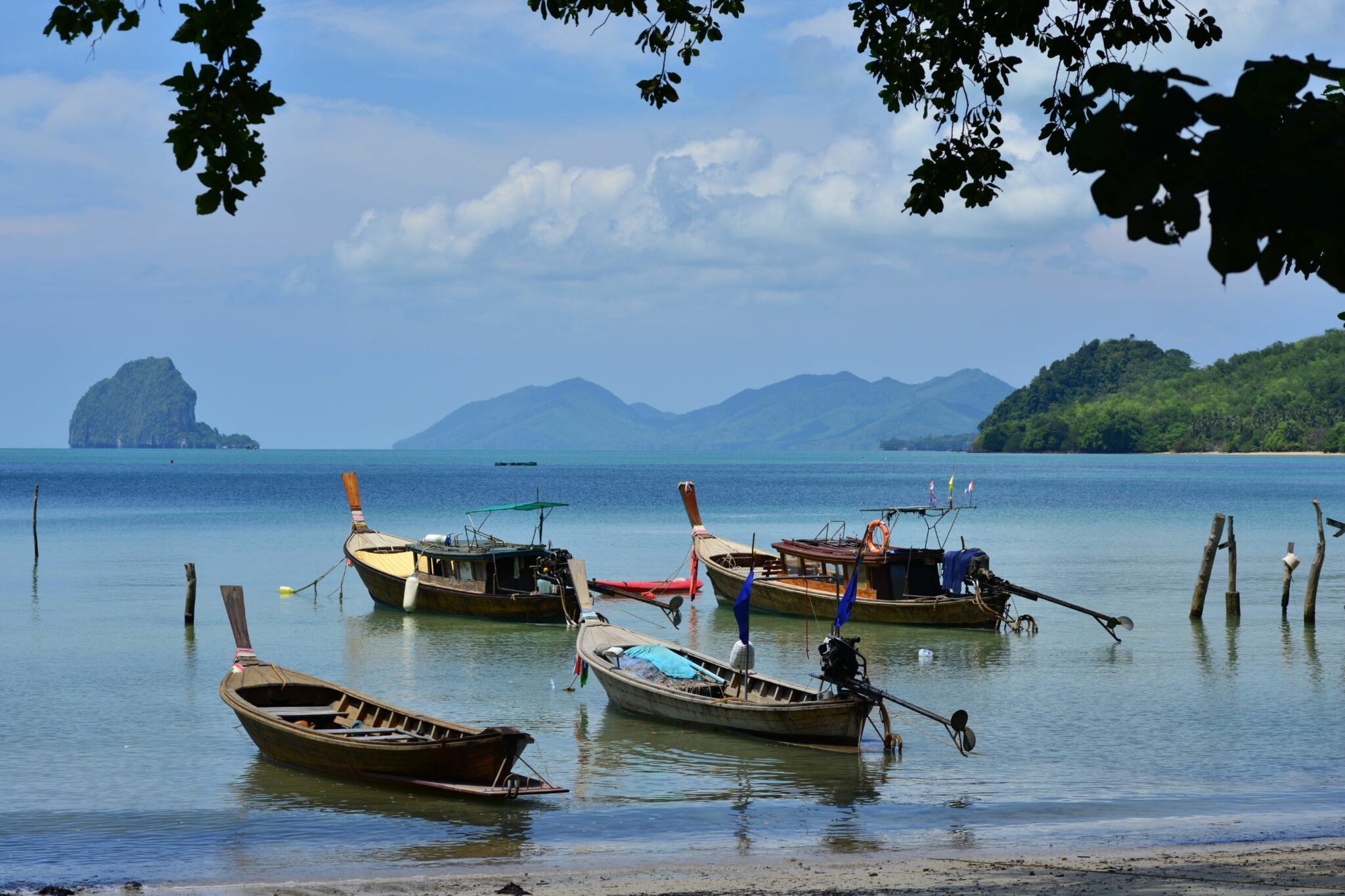 Longtail boats float in shallow water near a quiet beach, with distant islands and hazy mountains on the horizon.