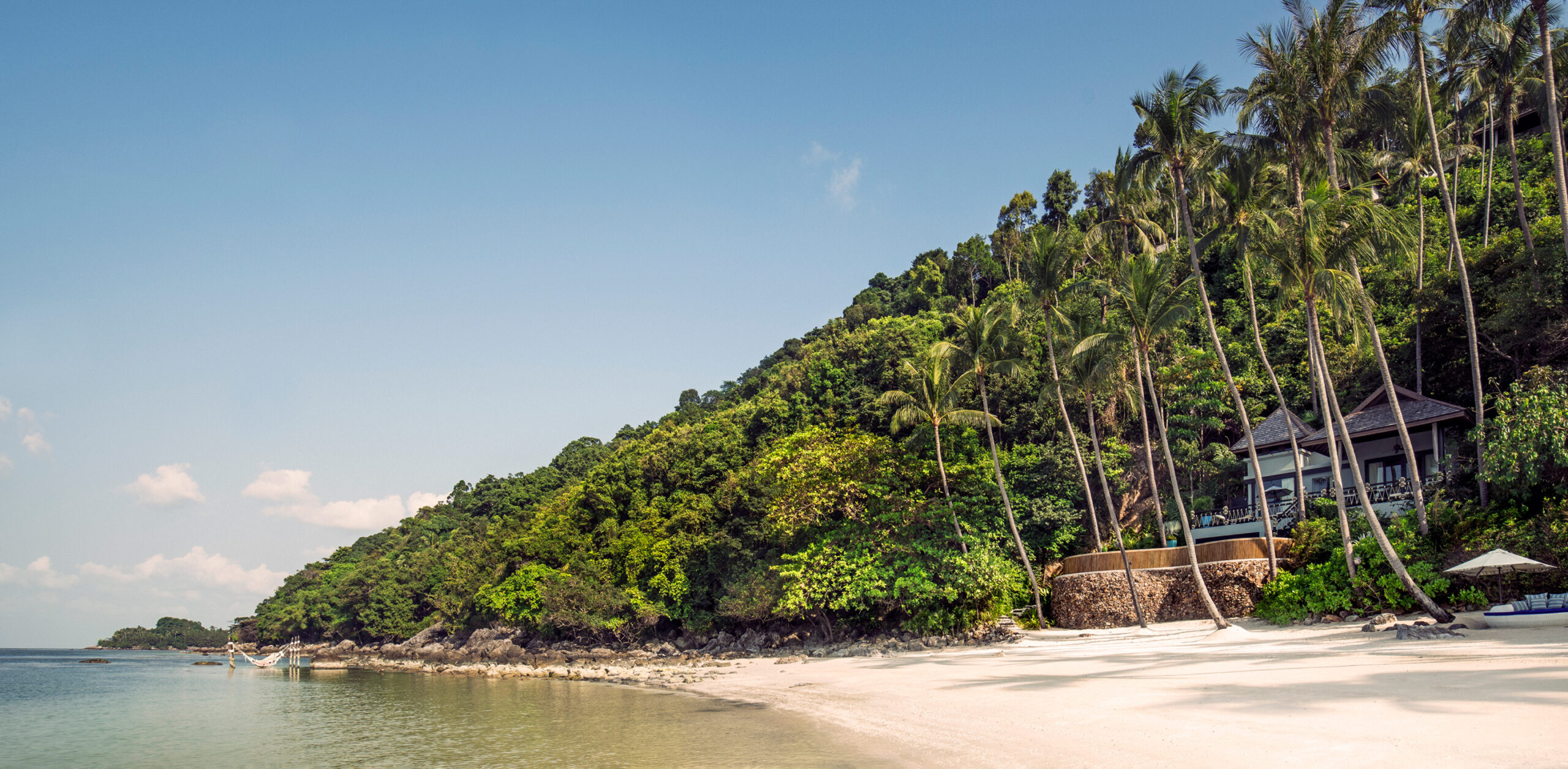 A palm-lined beach meets clear shallow water, with villas tucked into a lush hillside under a bright sky.