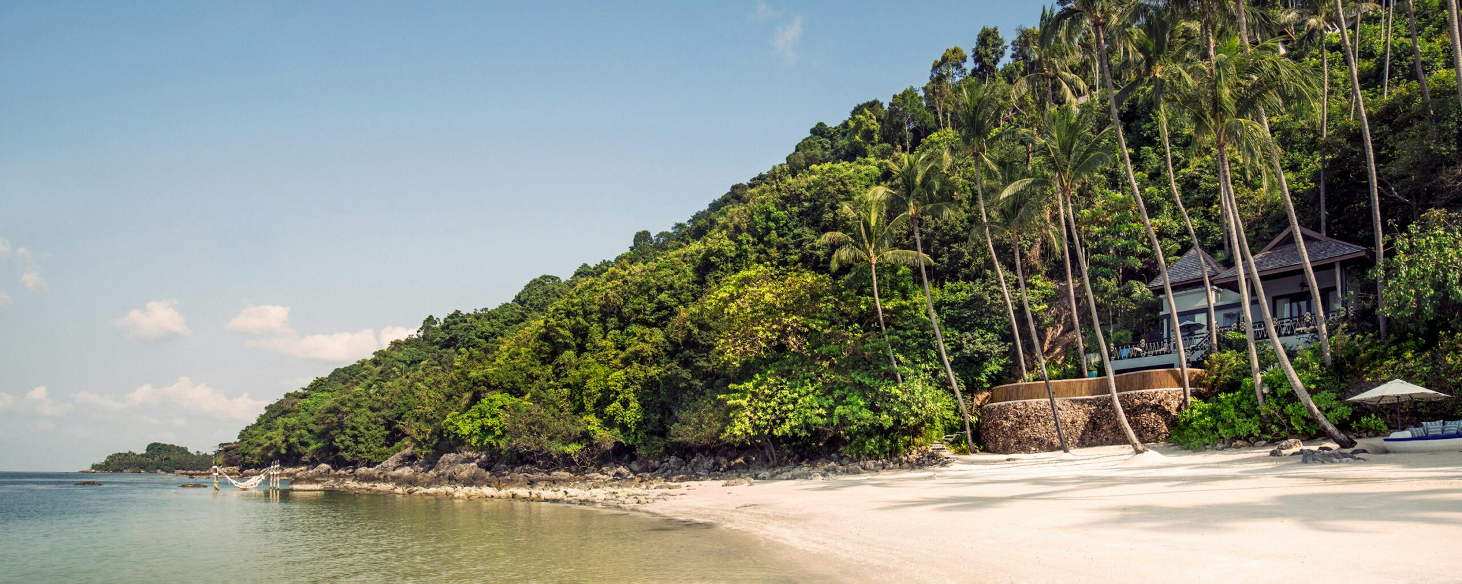 A palm-lined beach meets clear shallow water, with villas tucked into a lush hillside under a bright sky.