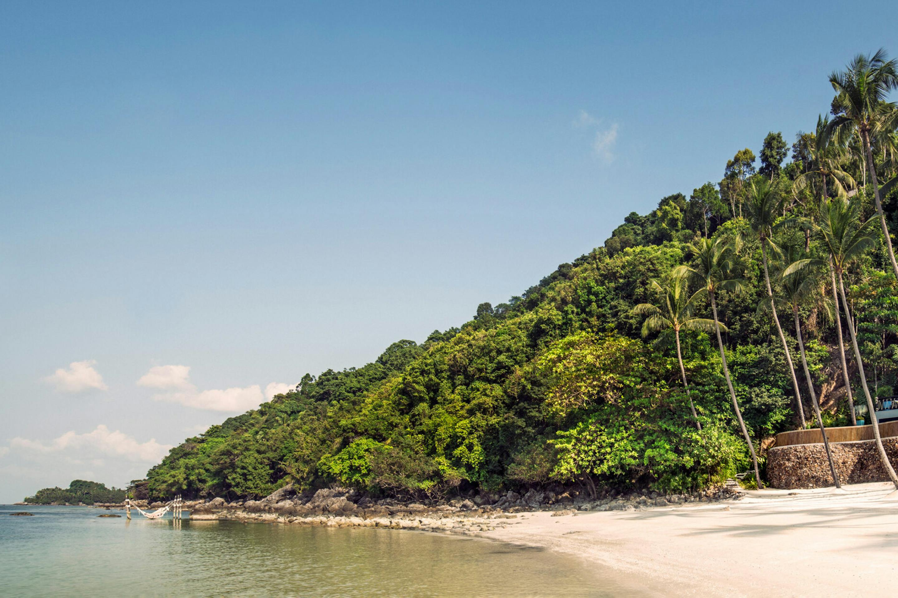 A palm-lined beach meets clear shallow water, with villas tucked into a lush hillside under a bright sky.
