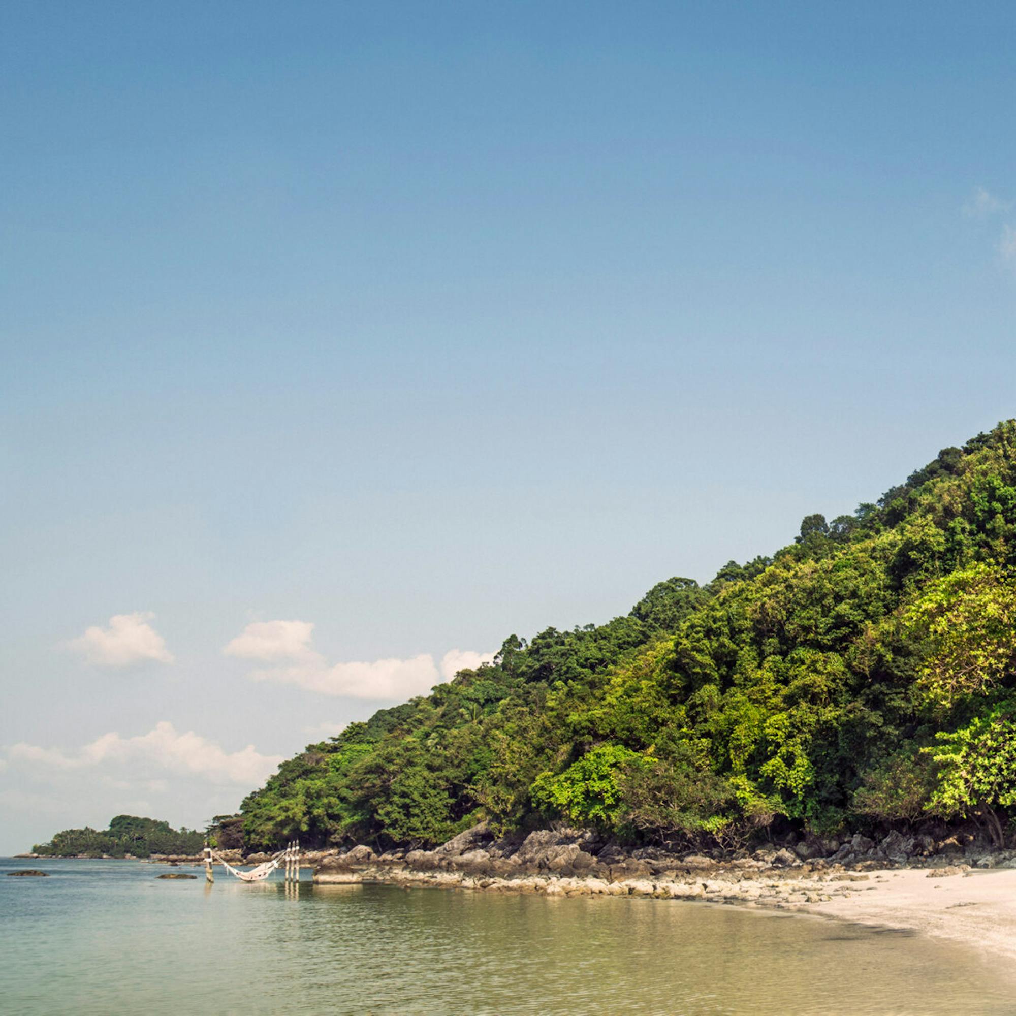 A palm-lined beach meets clear shallow water, with villas tucked into a lush hillside under a bright sky.