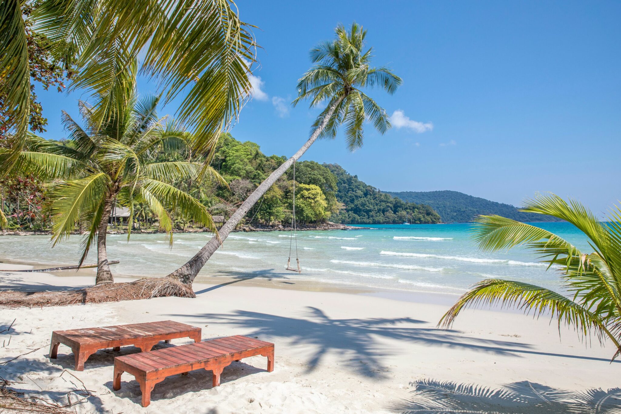Wooden loungers sit on white sand beneath palm trees, facing turquoise waves rolling into a sheltered bay.