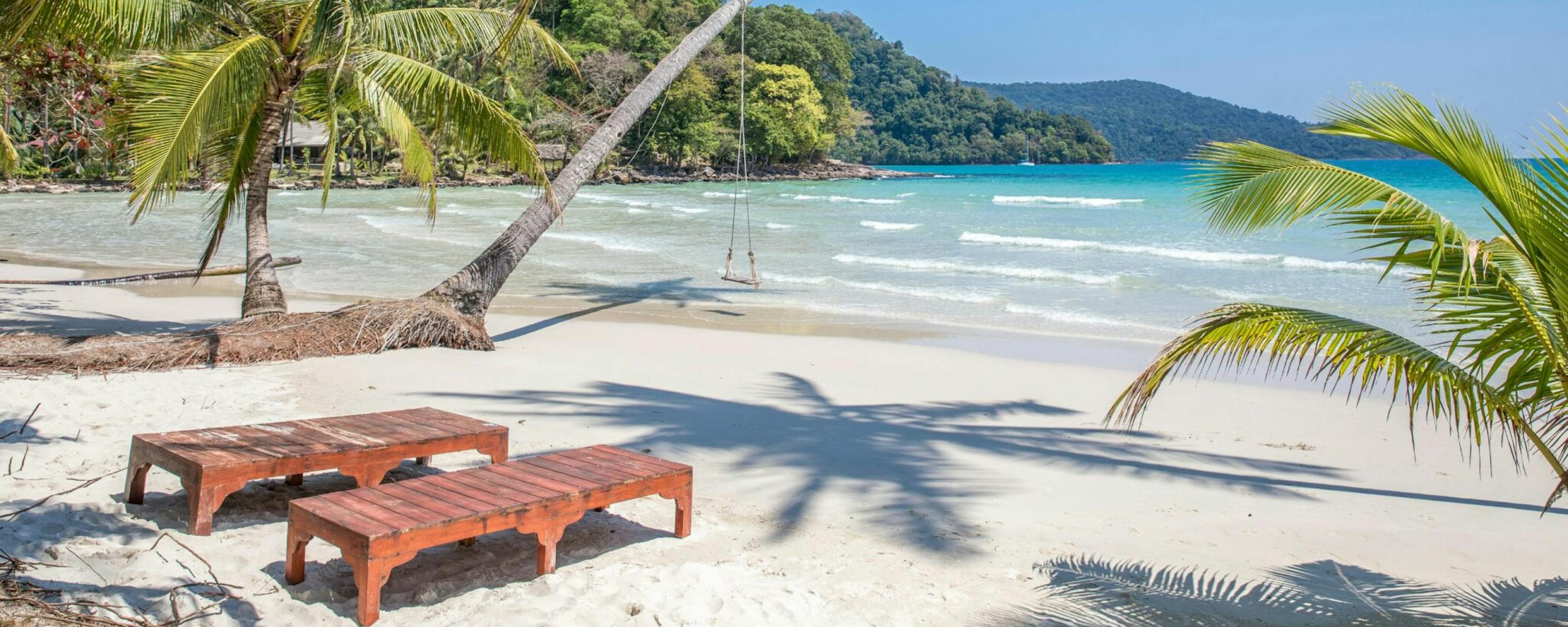 Wooden loungers sit on white sand beneath palm trees, facing turquoise waves rolling into a sheltered bay.