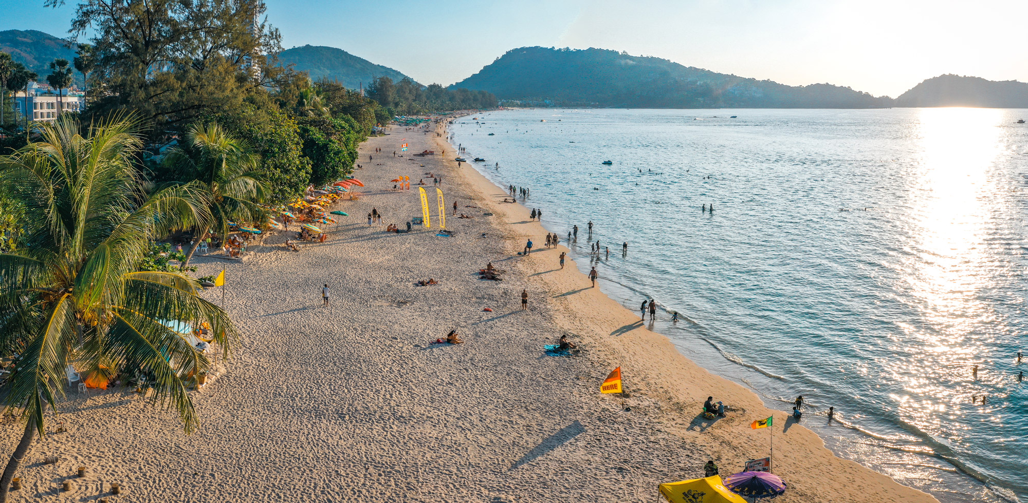 A long sandy beach curves beside calm water, dotted with umbrellas and people, with low hills in the distance.
