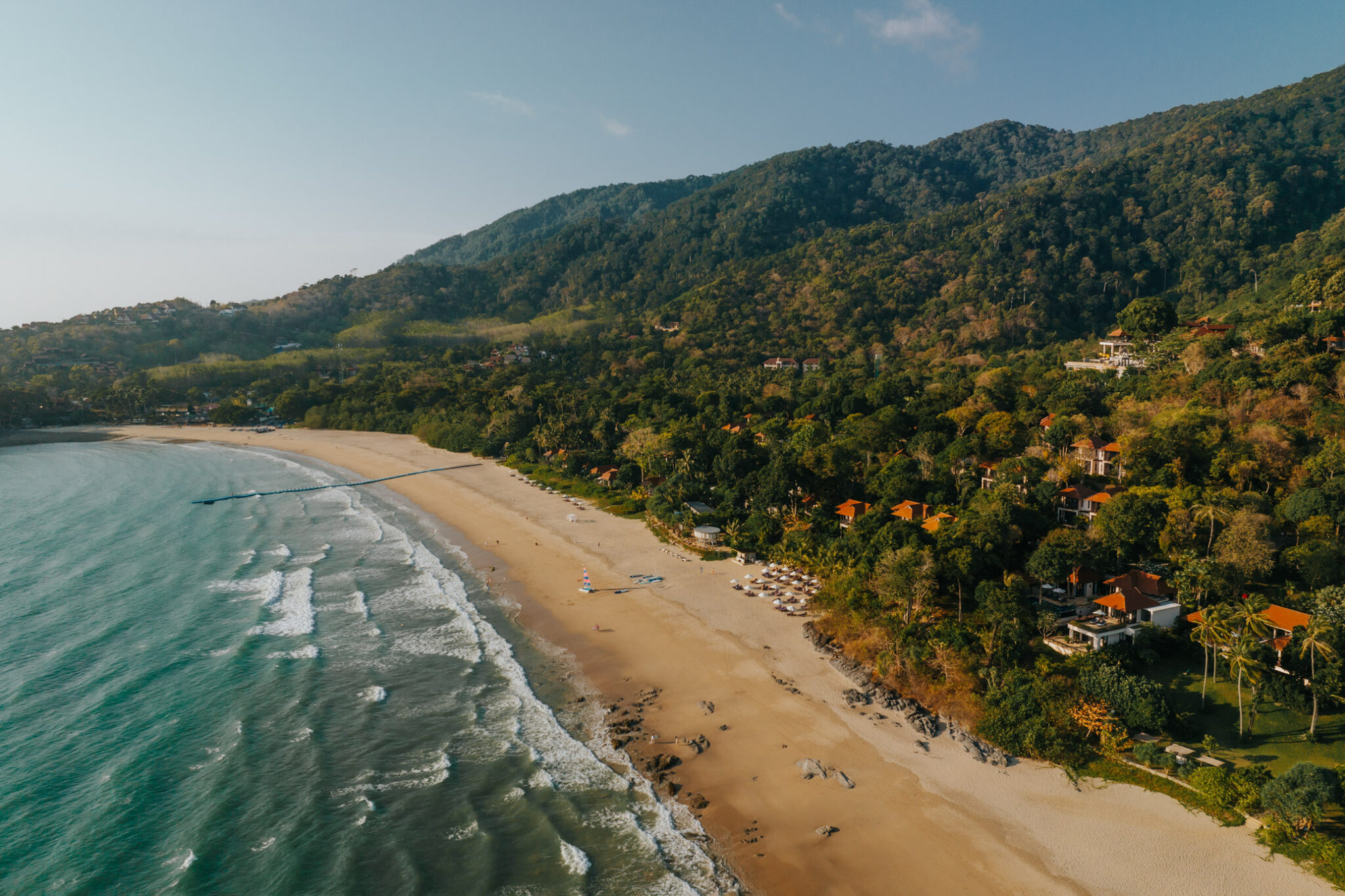 An aerial view shows a long beach and gentle surf, with dense forest and resort buildings along the shoreline.