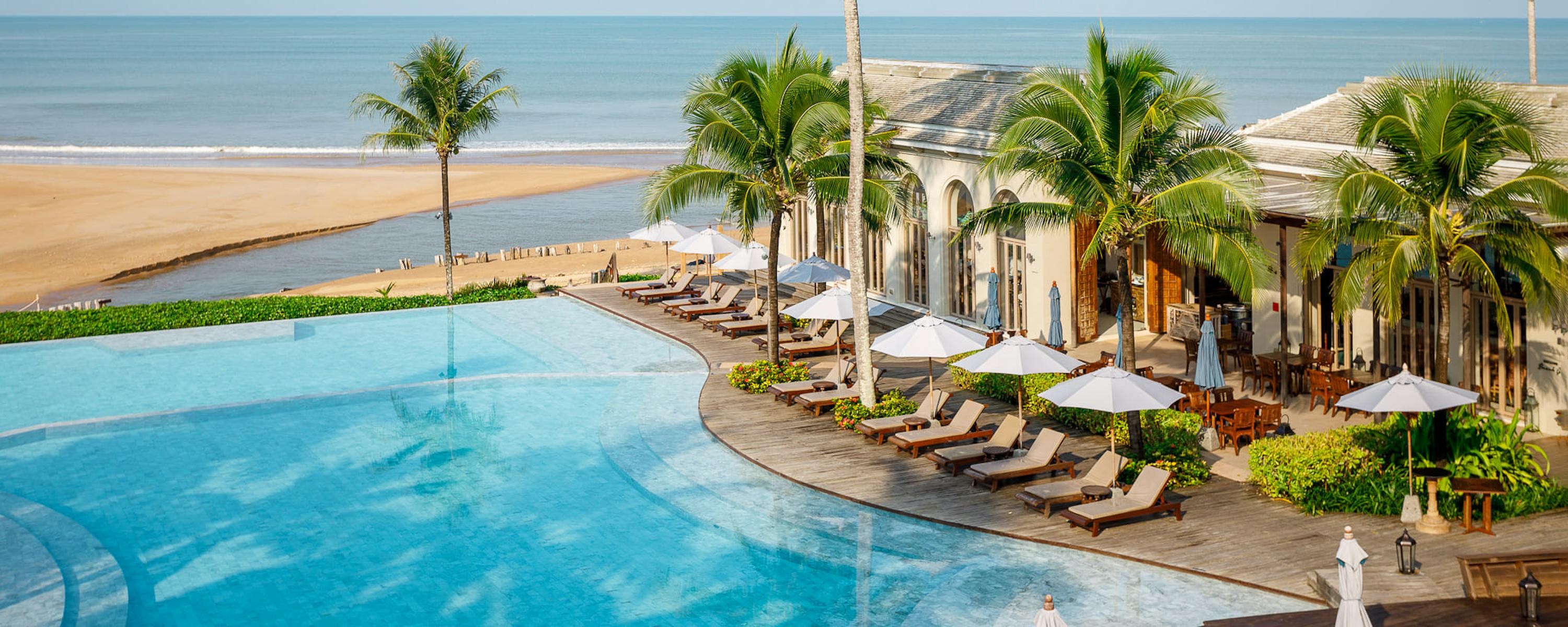 An infinity pool curves beside palms and sun loungers, facing a wide sandy beach and calm sea under clear light.