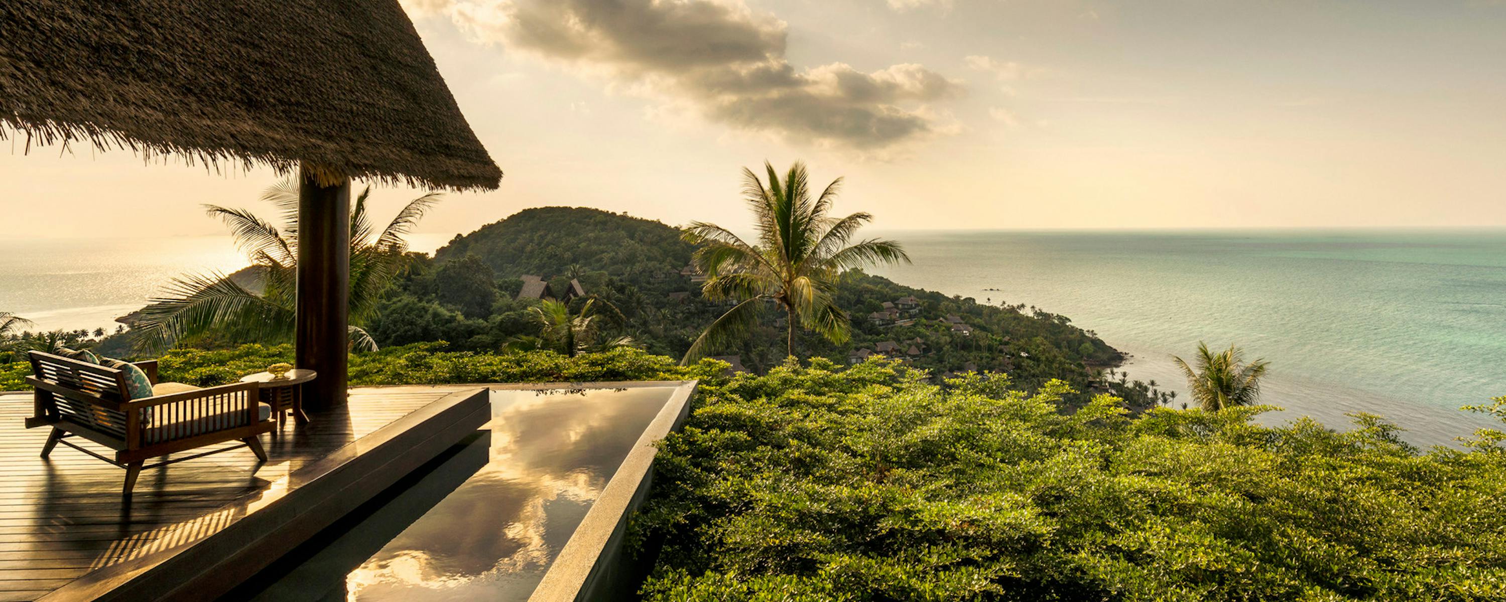 A thatched-roof terrace overlooks green hills and the sea, with lounge chairs set beside a reflective ledge.