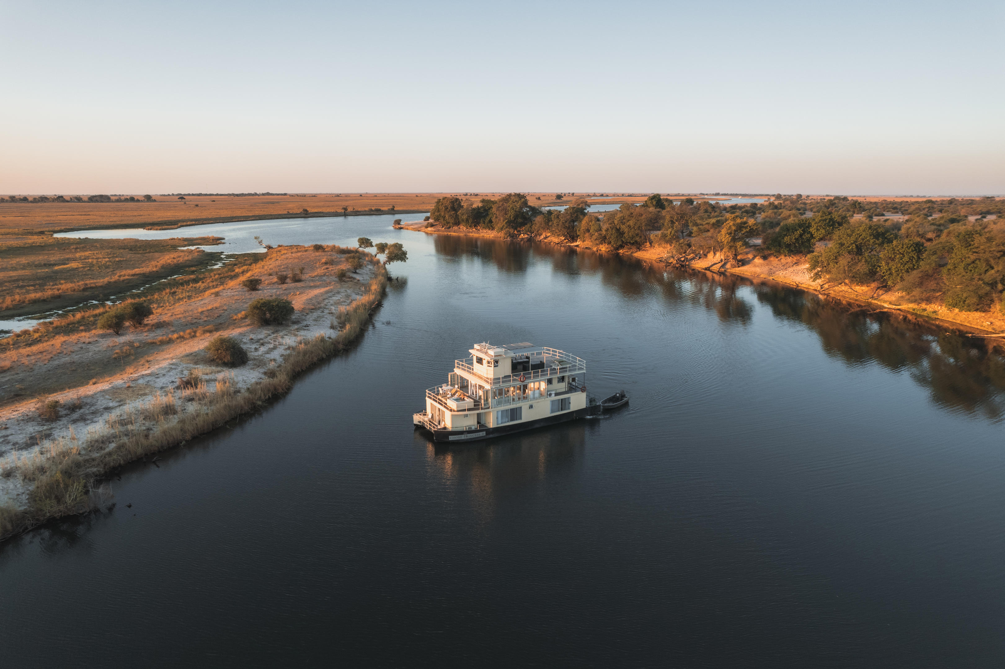 A river cruiser moves along the Chobe River at sunset, framed by sandbars and forested banks in an aerial view.