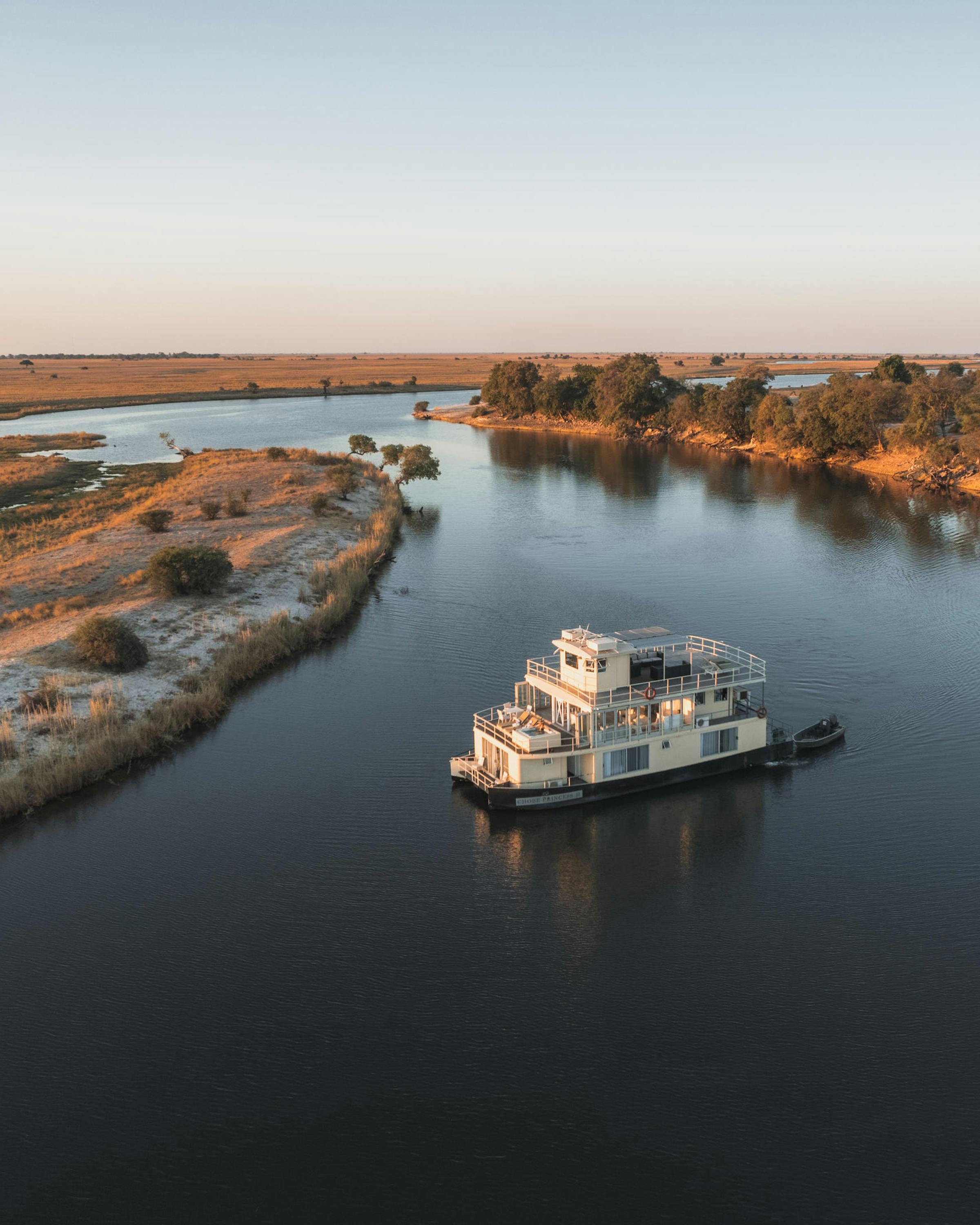 A river cruiser moves along the Chobe River at sunset, framed by sandbars and forested banks in an aerial view.