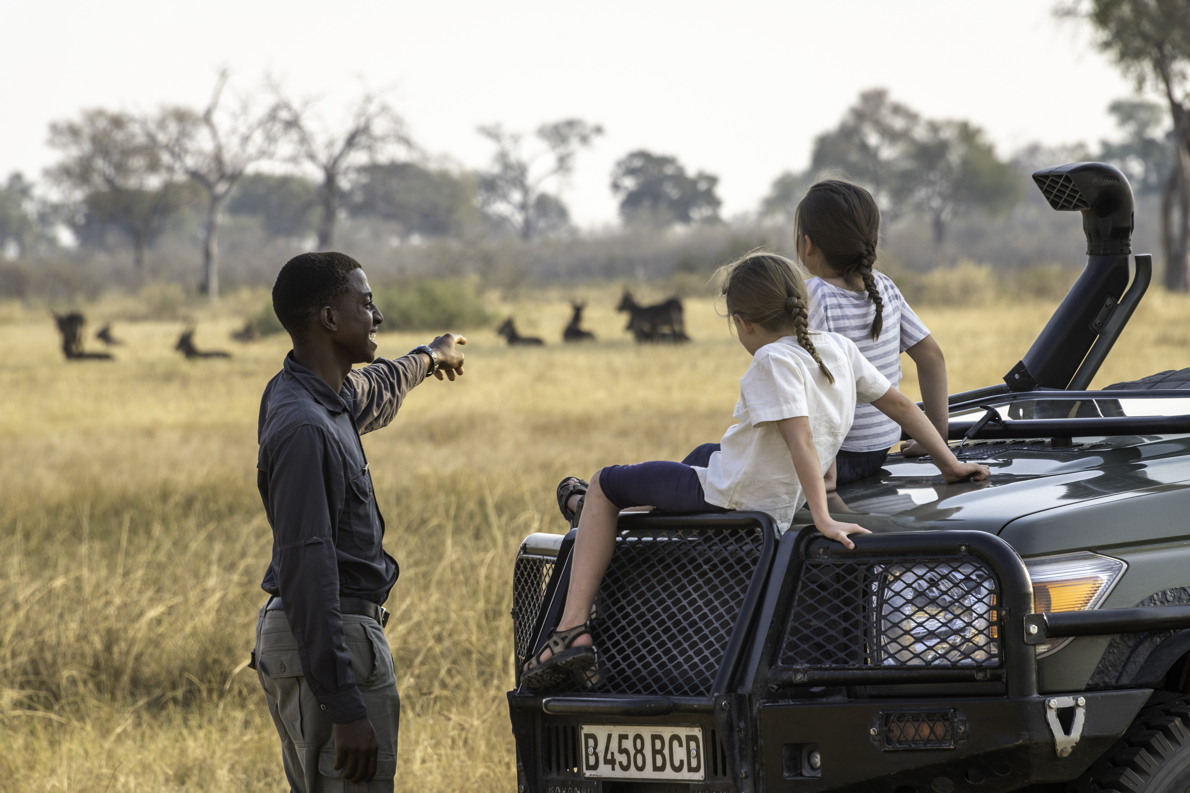 A guide chats with two children seated on a safari vehicle while herds graze across the dry plain behind.
