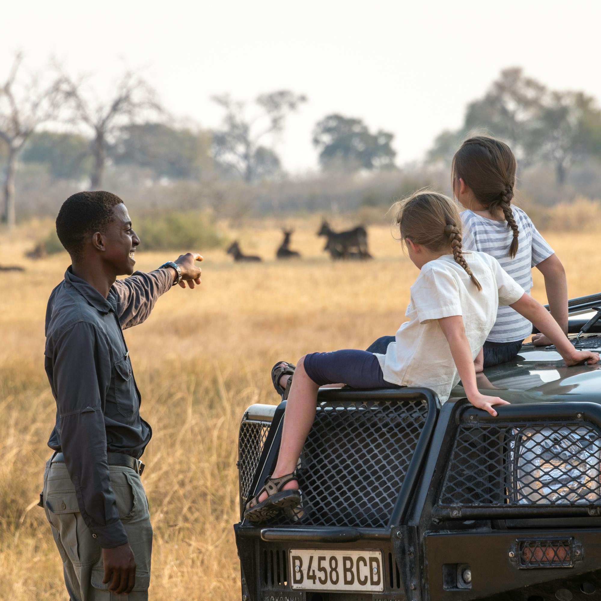 A guide chats with two children seated on a safari vehicle while herds graze across the dry plain behind.
