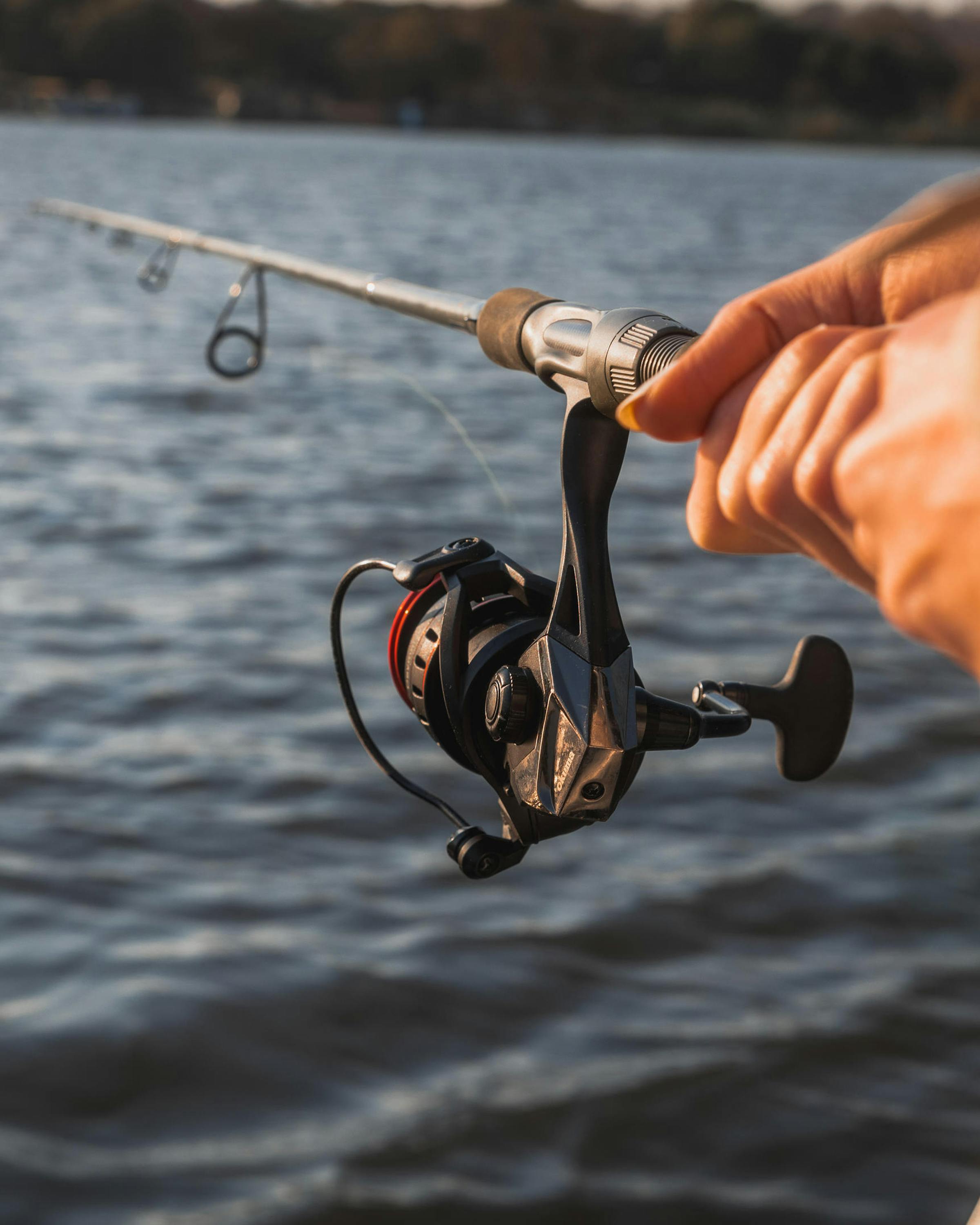 A hand holds a fishing rod over rippling river water, reel and line taut as the lure skims near the surface.