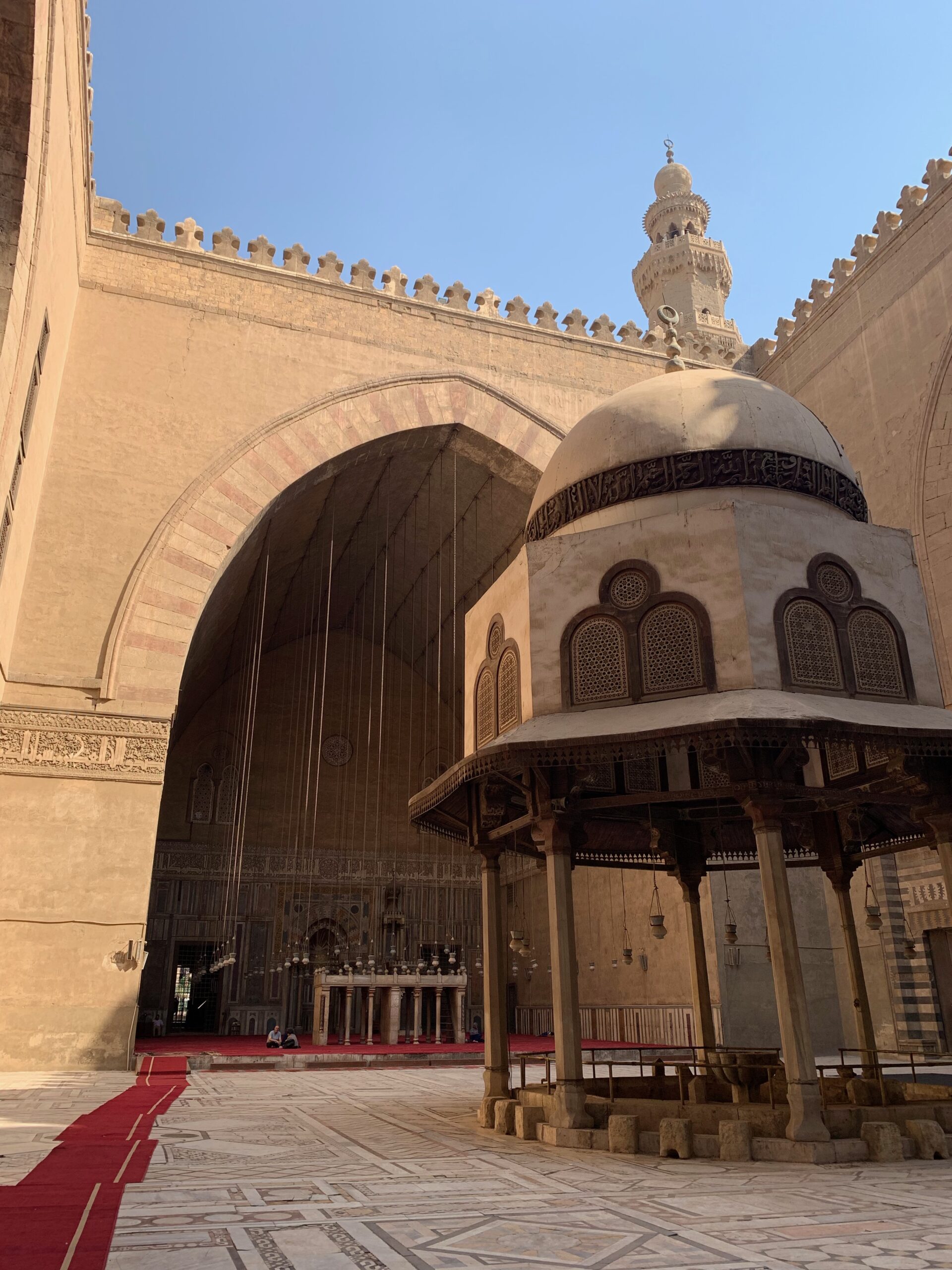 Arched colonnades frame a mosque courtyard, with a domed canopy and patterned stonework lit by afternoon sun.