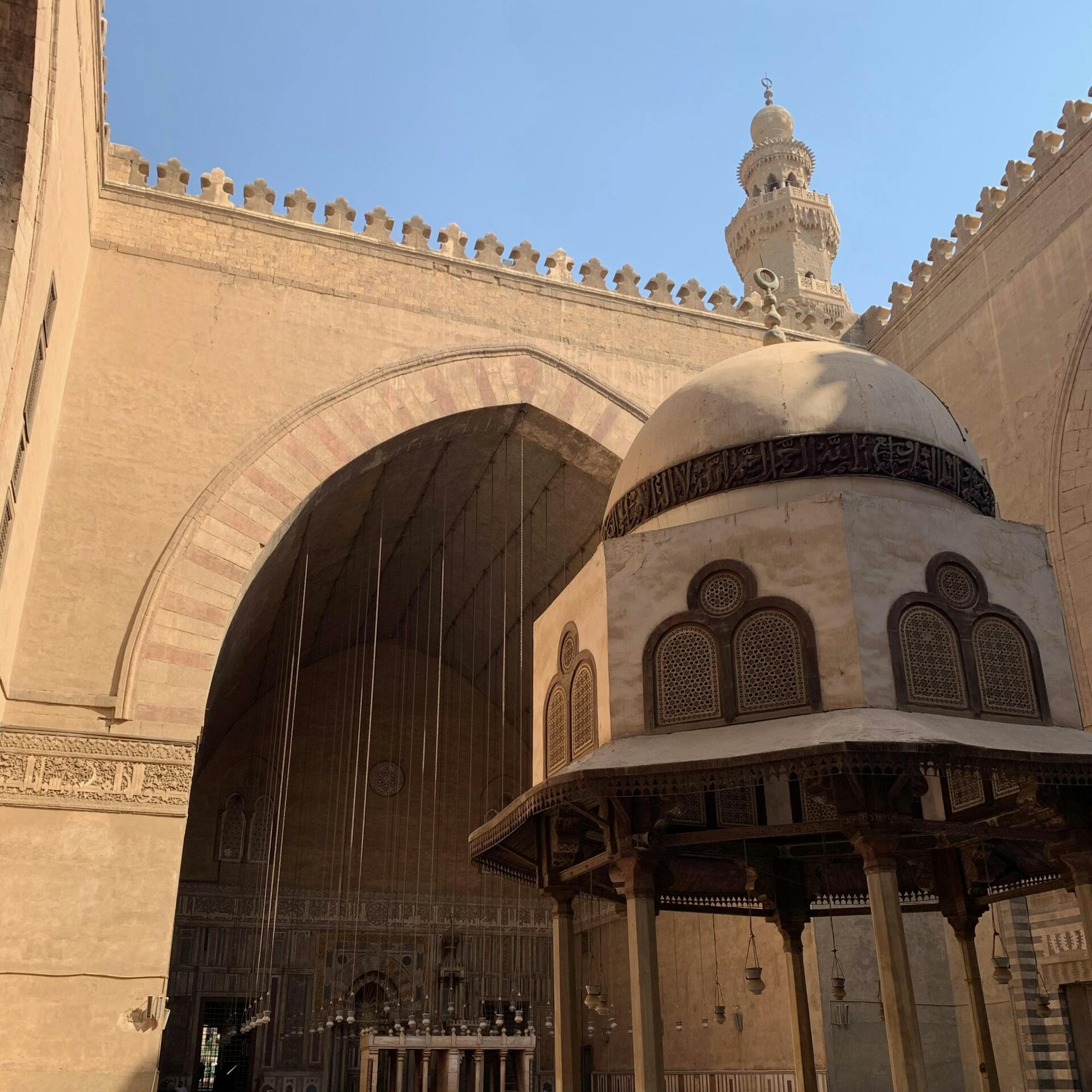 Arched colonnades frame a mosque courtyard, with a domed canopy and patterned stonework lit by afternoon sun.