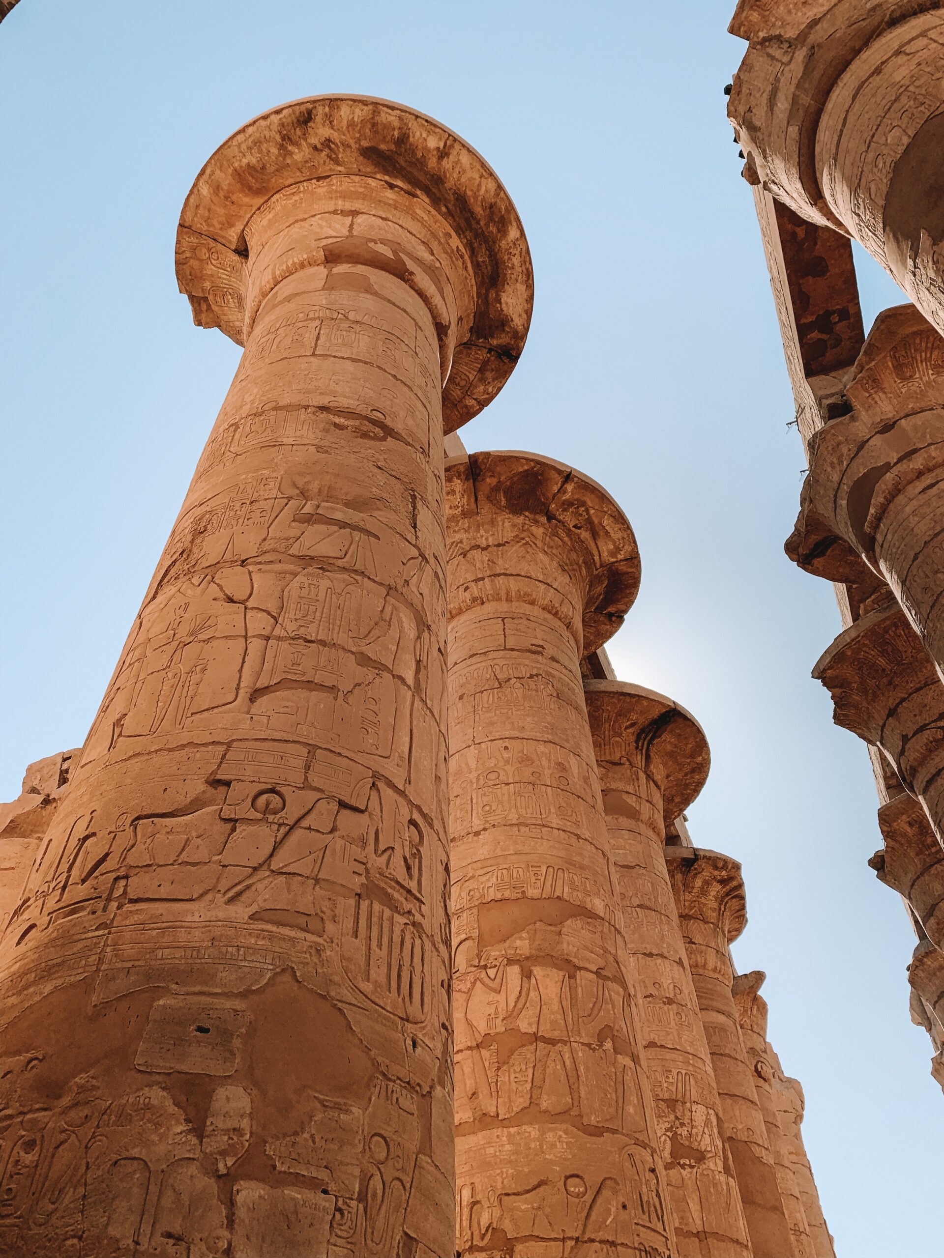 Upward view of a towering Karnak temple column carved with hieroglyphs, framed by warm stone and blue sky.