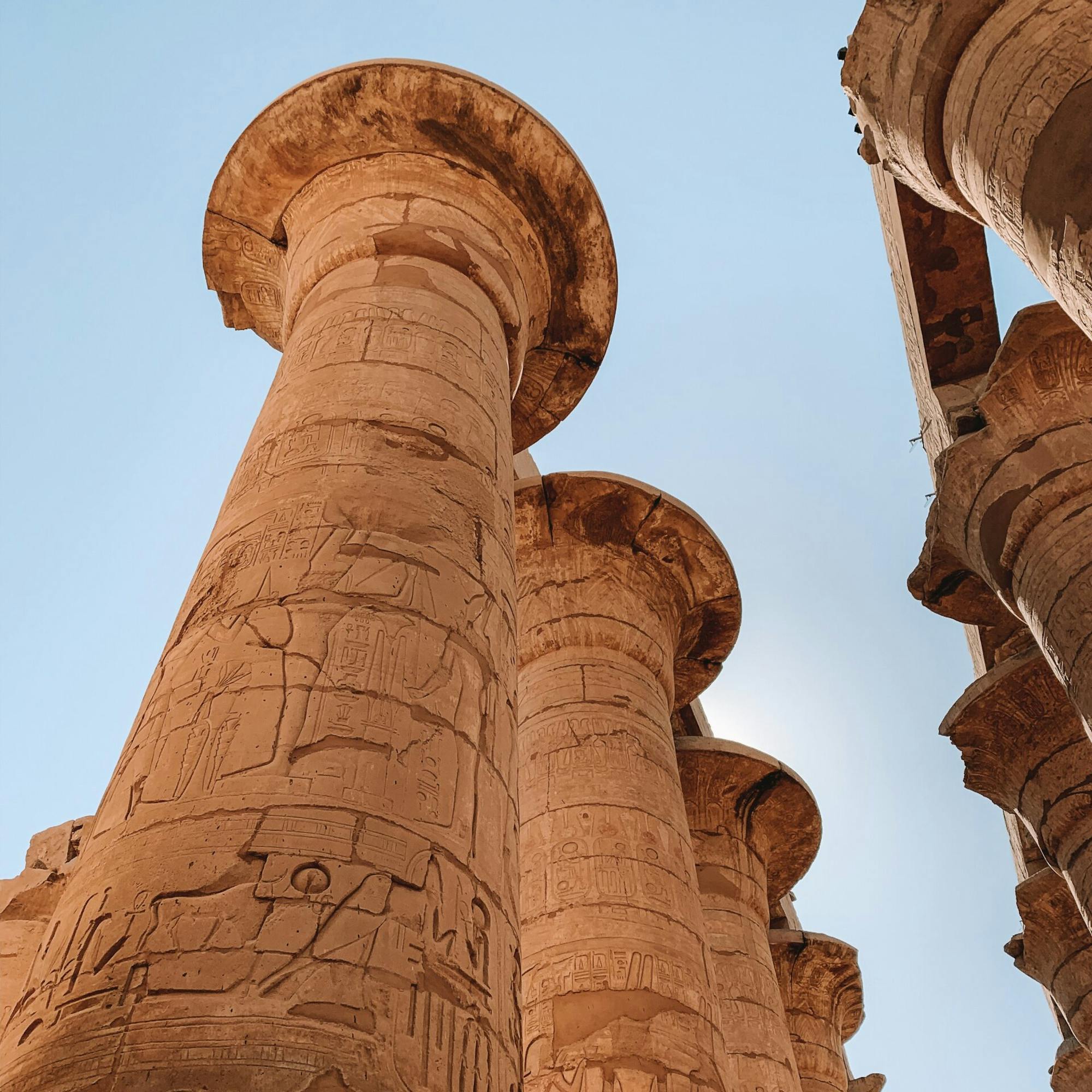 Upward view of a towering Karnak temple column carved with hieroglyphs, framed by warm stone and blue sky.