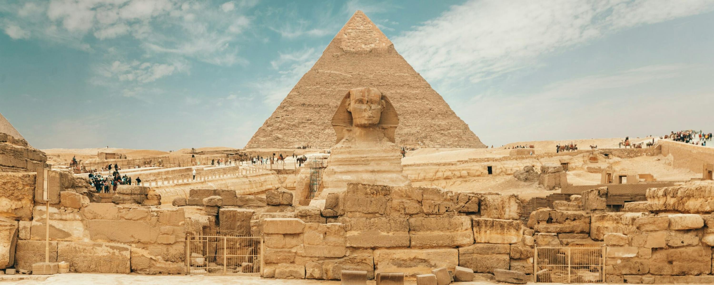 A traveler walks toward a large pyramid and ruins at Giza, with sandy stone blocks and a blue sky overhead.