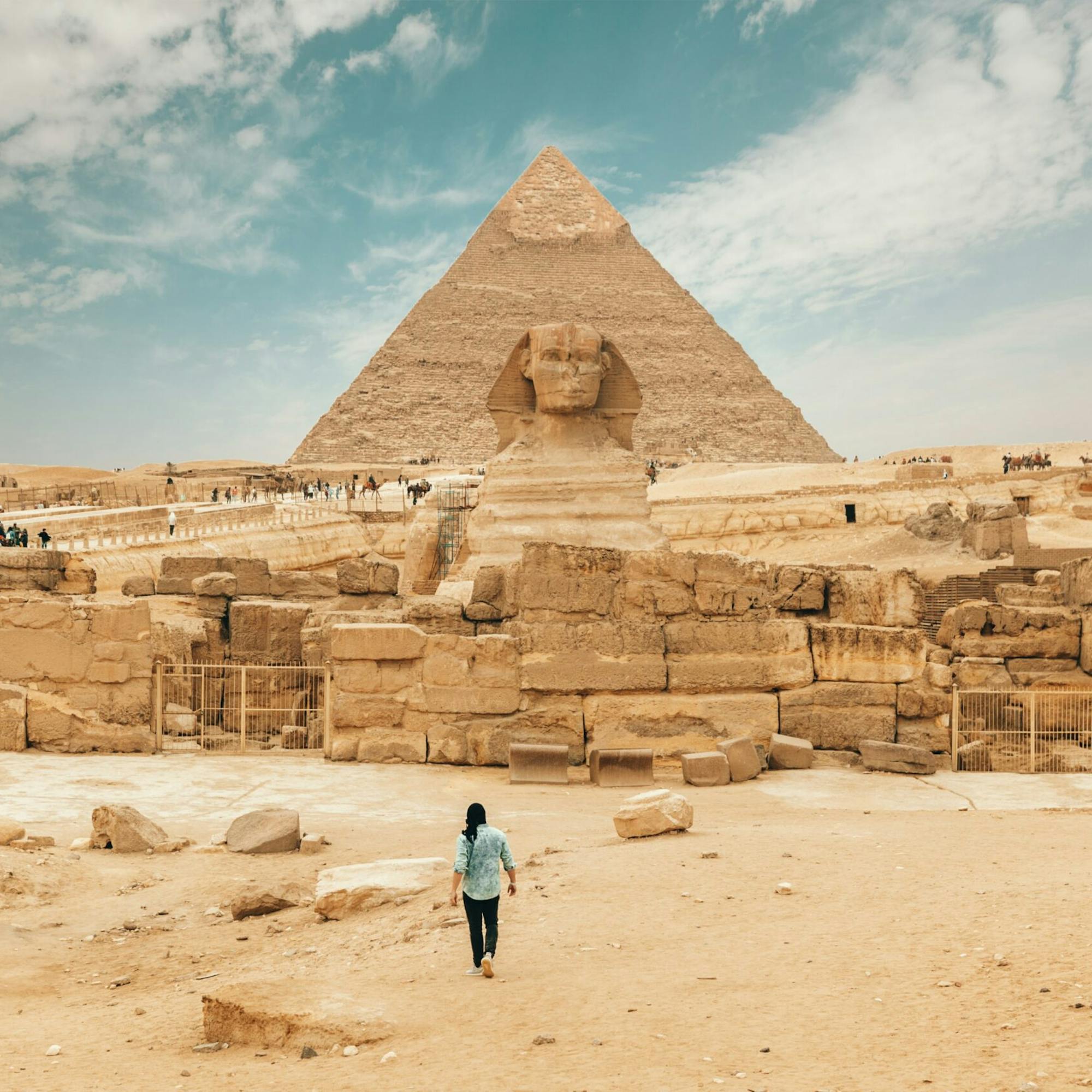 A traveler walks toward a large pyramid and ruins at Giza, with sandy stone blocks and a blue sky overhead.