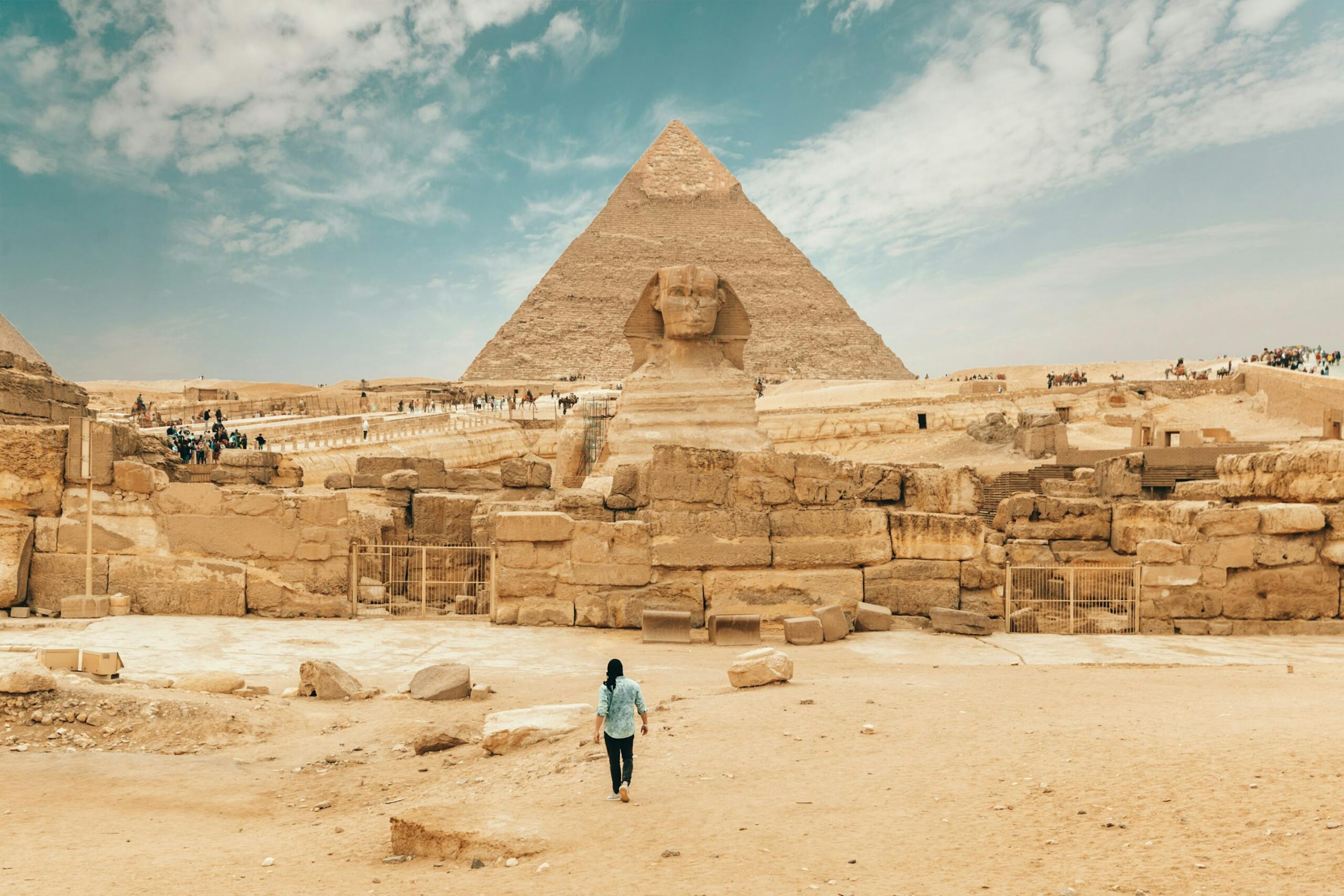 A traveler walks toward a large pyramid and ruins at Giza, with sandy stone blocks and a blue sky overhead.