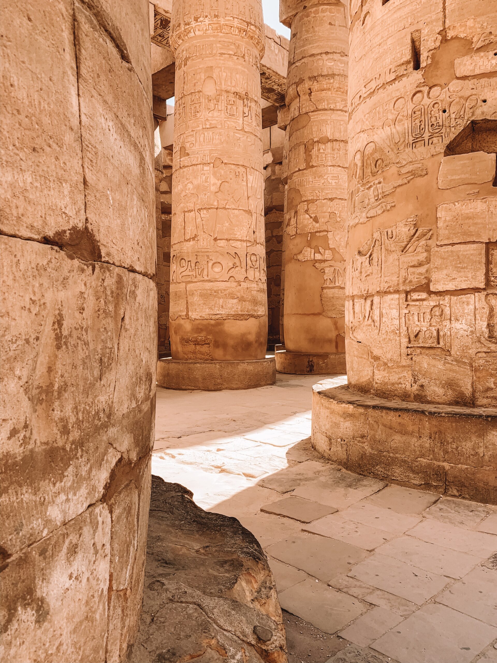 Sunlit sandstone columns and passageways at Karnak temple show worn carvings and shadows across the stone floor.
