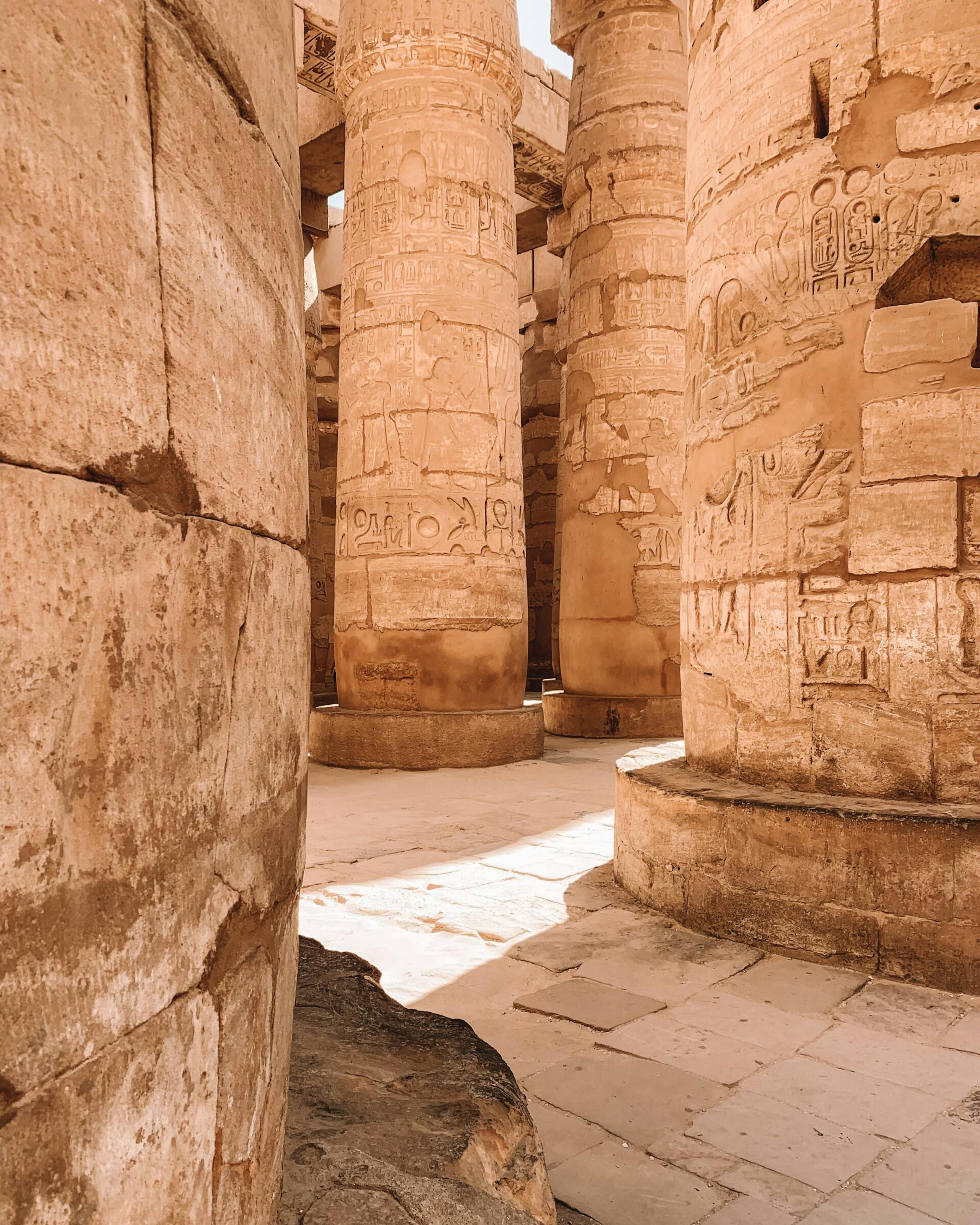 Sunlit sandstone columns and passageways at Karnak temple show worn carvings and shadows across the stone floor.
