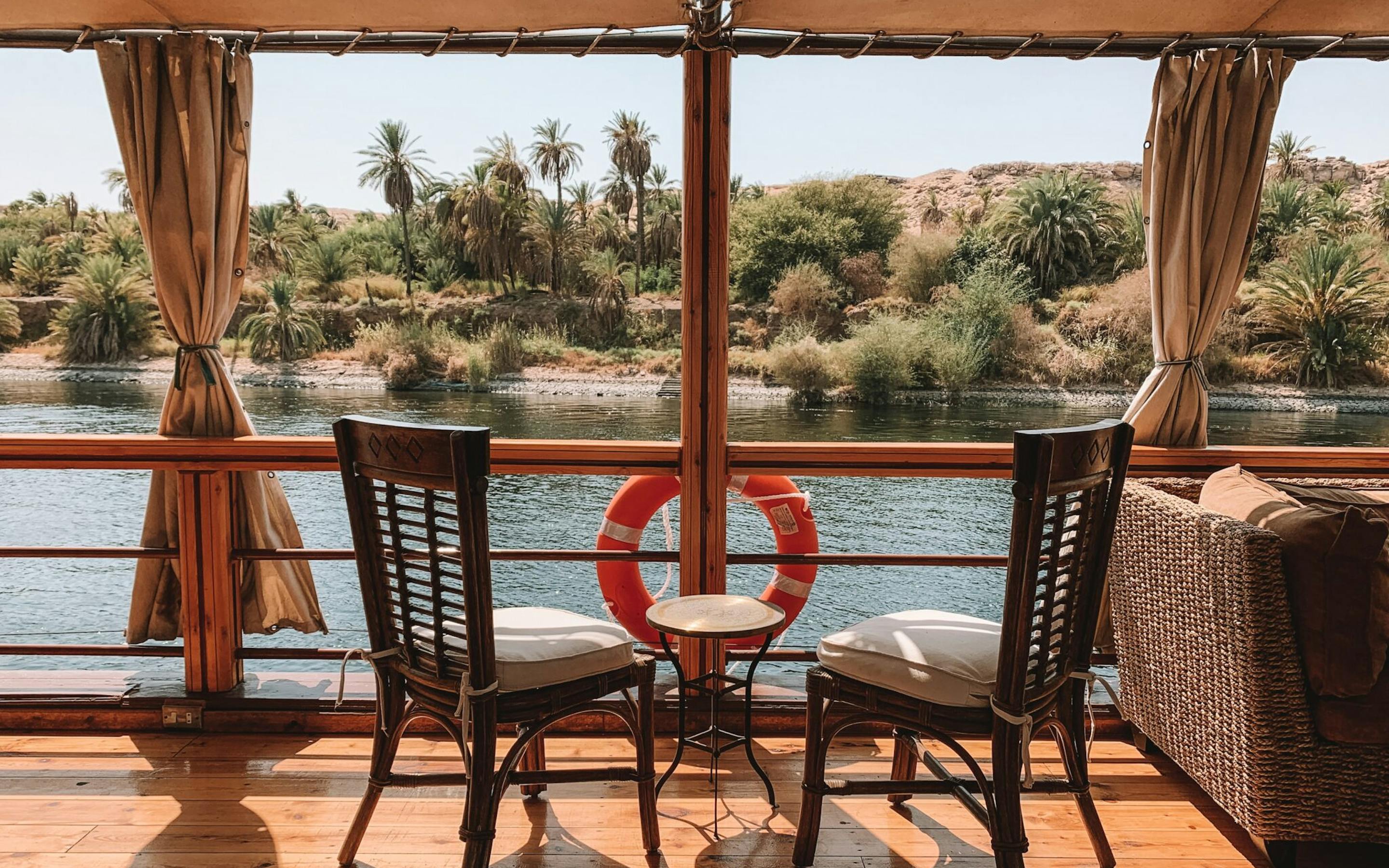 Two wooden chairs and a life ring sit on a shaded boat deck overlooking the Nile, with palms and water beyond.