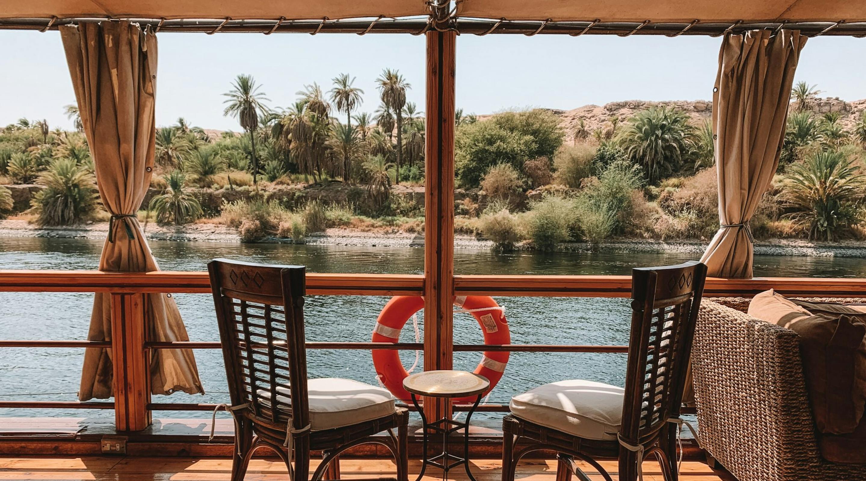 Two wooden chairs and a life ring sit on a shaded boat deck overlooking the Nile, with palms and water beyond.