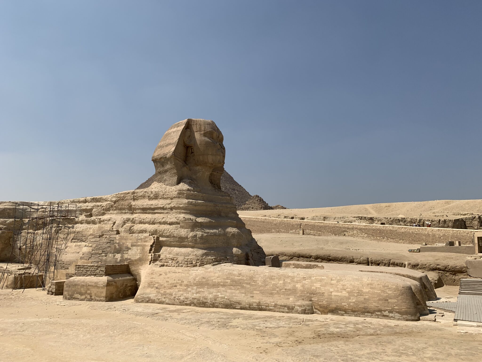 The Great Sphinx sits in a sandy desert plain under clear skies, with low stone walls and worn steps nearby.