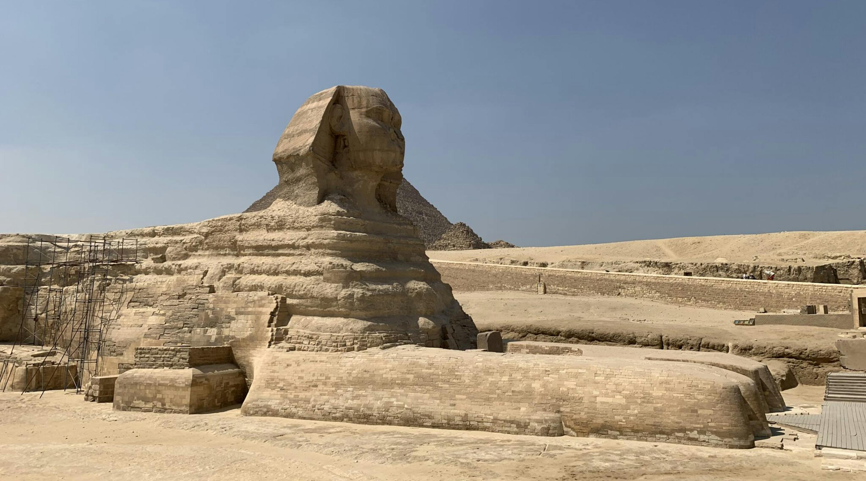 The Great Sphinx sits in a sandy desert plain under clear skies, with low stone walls and worn steps nearby.