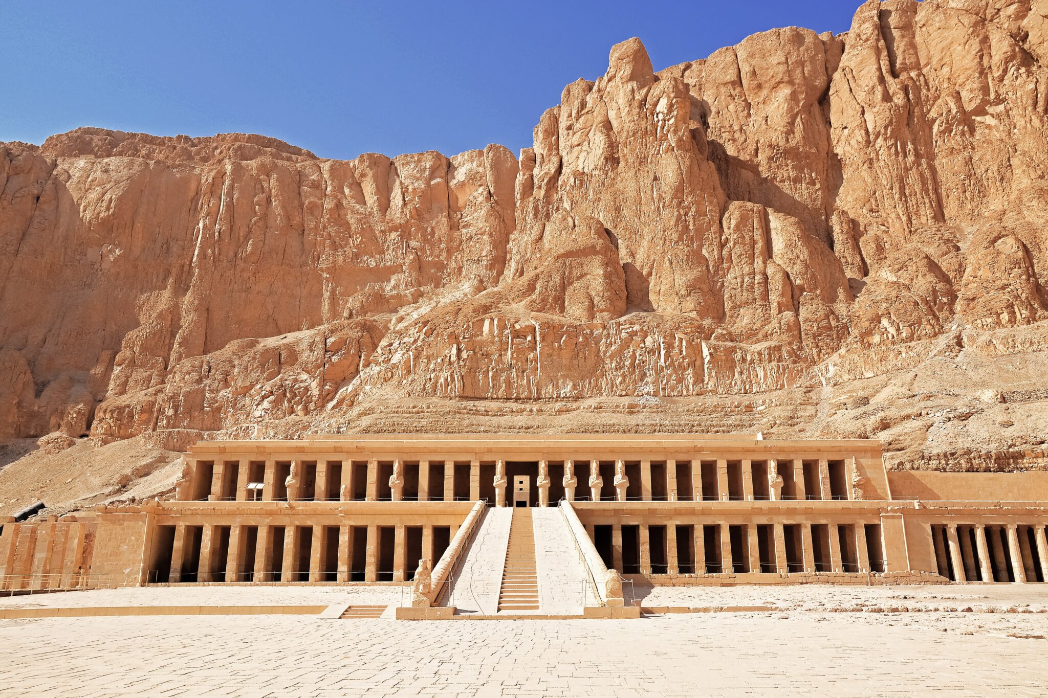 A terraced stone temple sits at the base of towering desert cliffs, with a central ramp leading to colonnades.