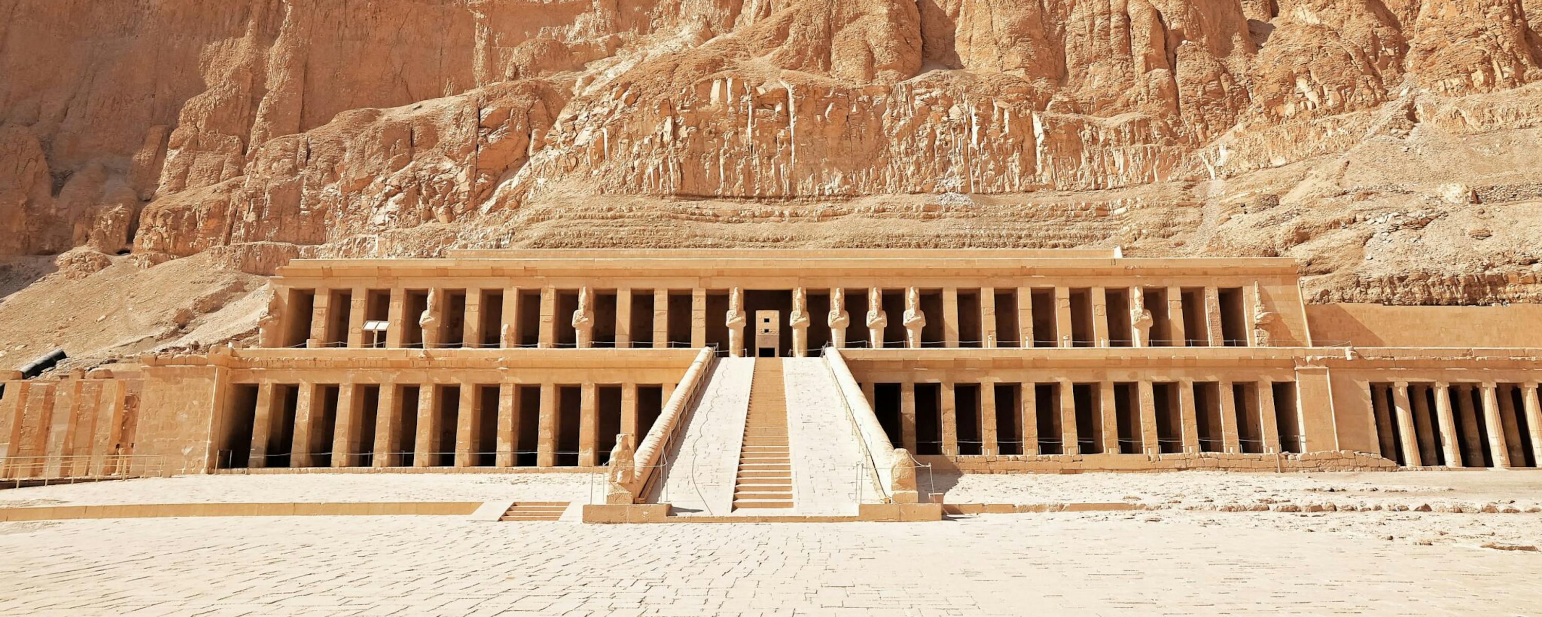 A terraced stone temple sits at the base of towering desert cliffs, with a central ramp leading to colonnades.