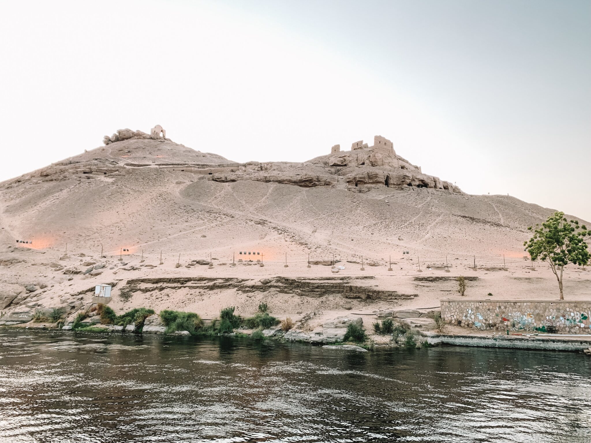 Low desert hills and small buildings sit across the Nile near Aswan, reflected in rippling water under pale light.